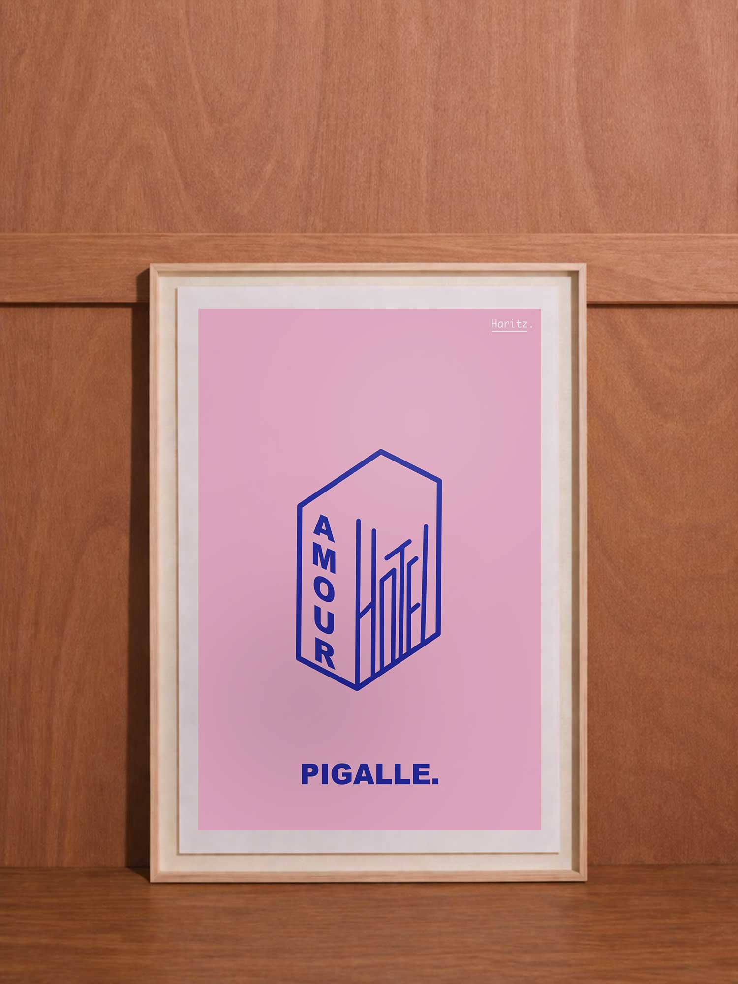Affiche contemporaine de décoration Haritz sur fond rose dégradé représentant l'Hôtel Amour de Pigalle avec illustration d'un bâtiment en perspective isométrique bleu marine portant le mot "AMOUR" verticalement, encadrée et posée contre un mur en bois