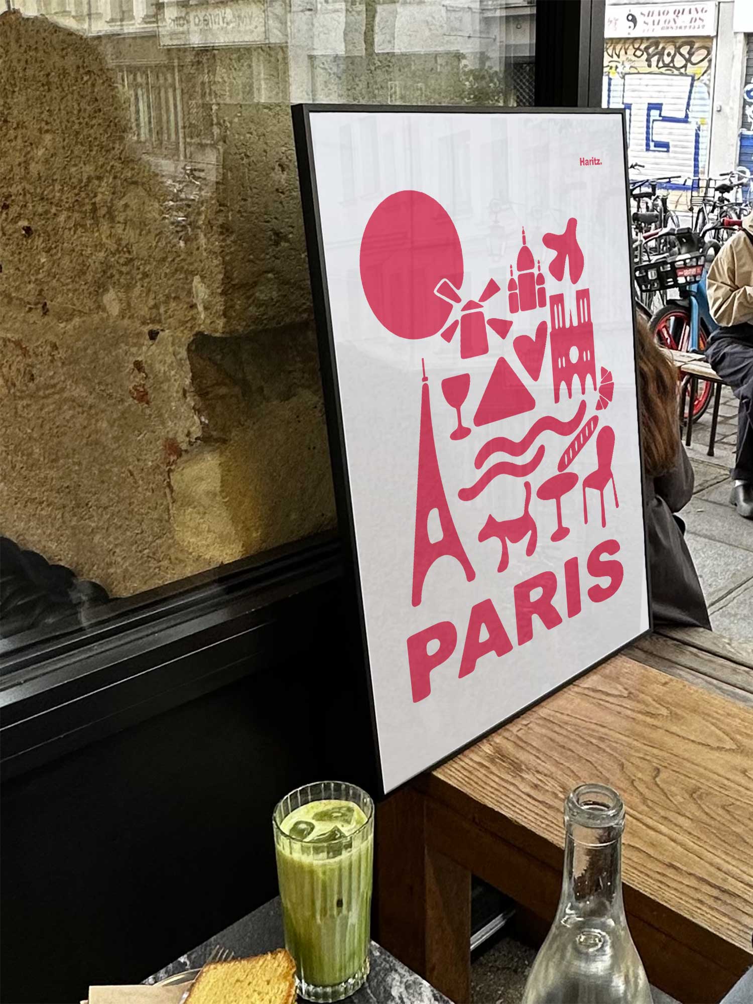 Affiche Paris, version rose et blanche, de Haritz exposée près d'une fenêtre dans un café parisien tendance à côté d'un verre de matcha latte.