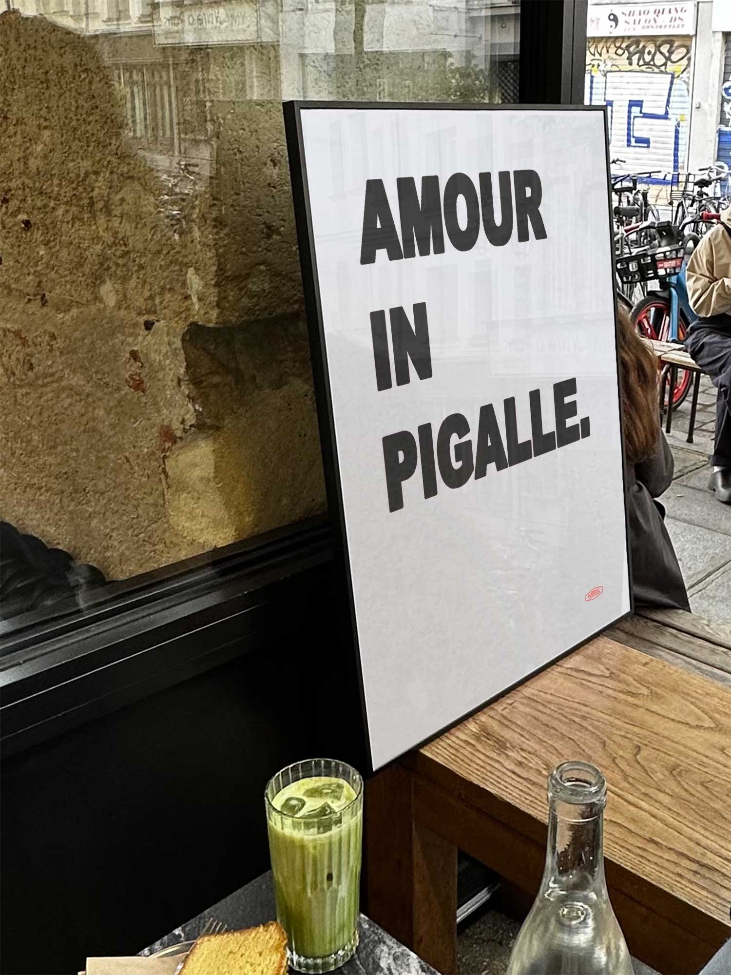 Affiche Amour In Pigalle de Haritz exposée près d'une fenêtre dans un café parisien tendance à côté d'un verre de matcha latte.