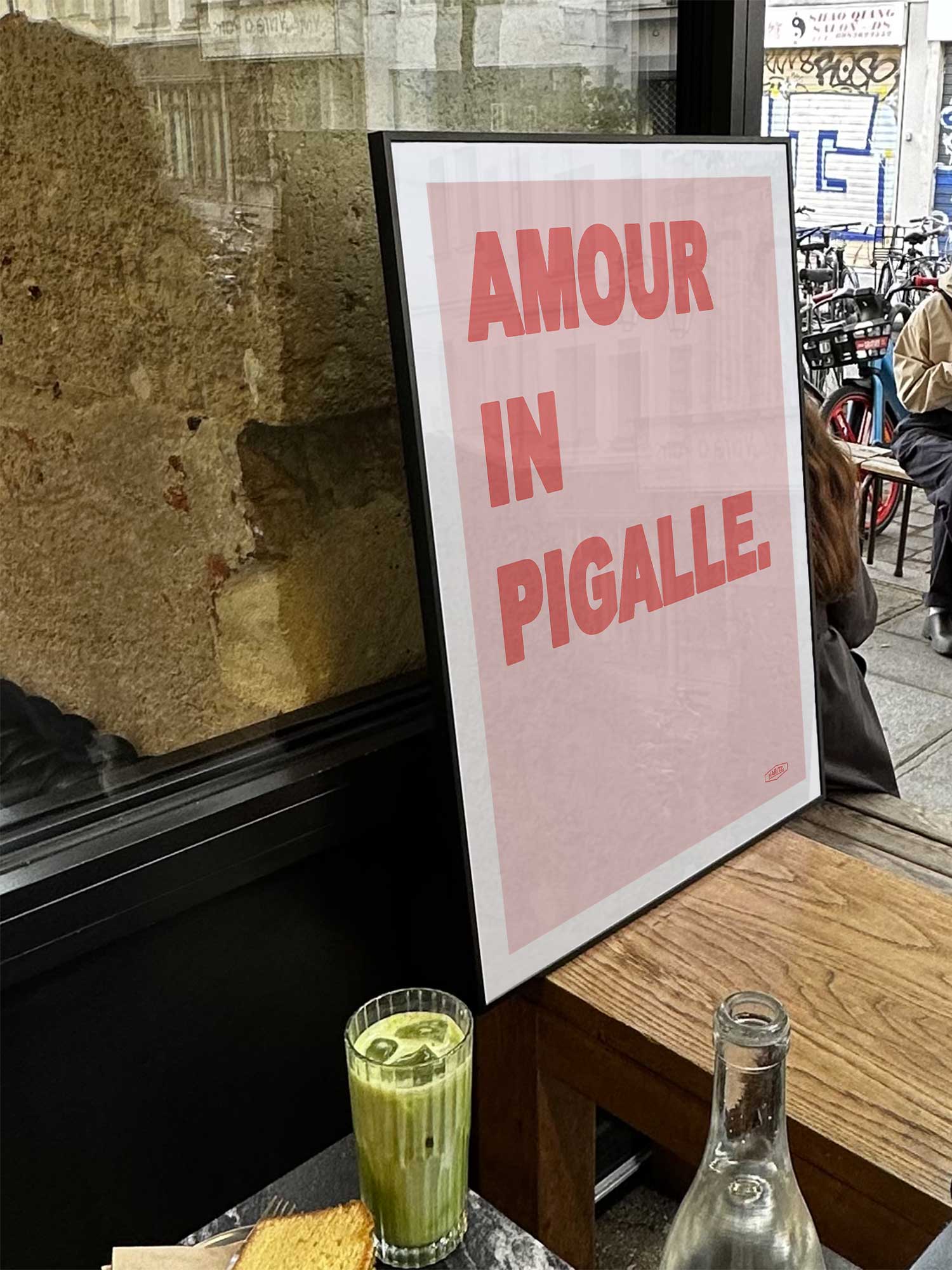 Affiche Amour in Pigalle de Haritz exposée près d'une fenêtre dans un café parisien tendance à côté d'un verre de matcha latte.