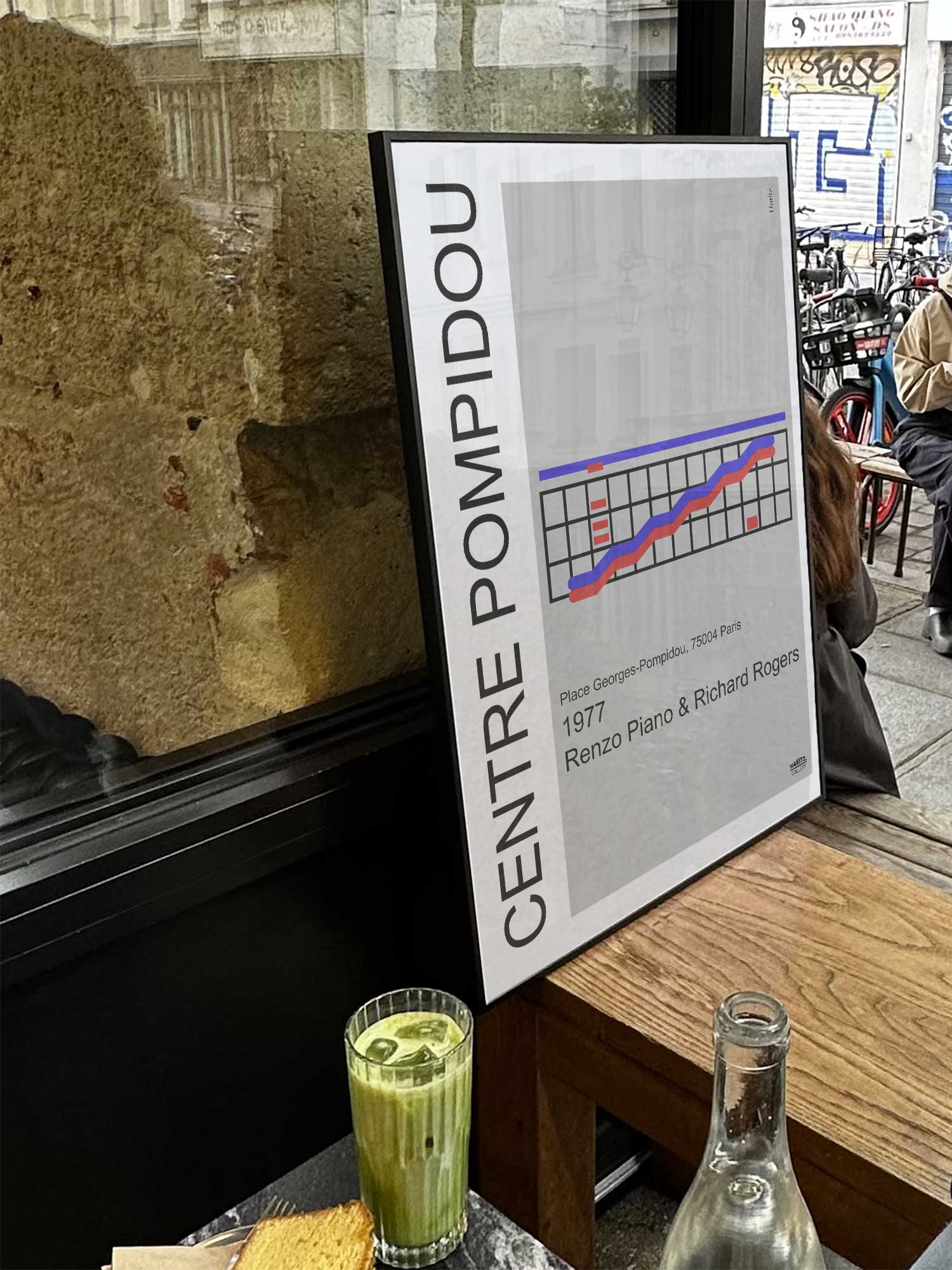 Affiche Centre Pompidou de Haritz exposée près d'une fenêtre dans un café parisien tendance à côté d'un verre de matcha latte.