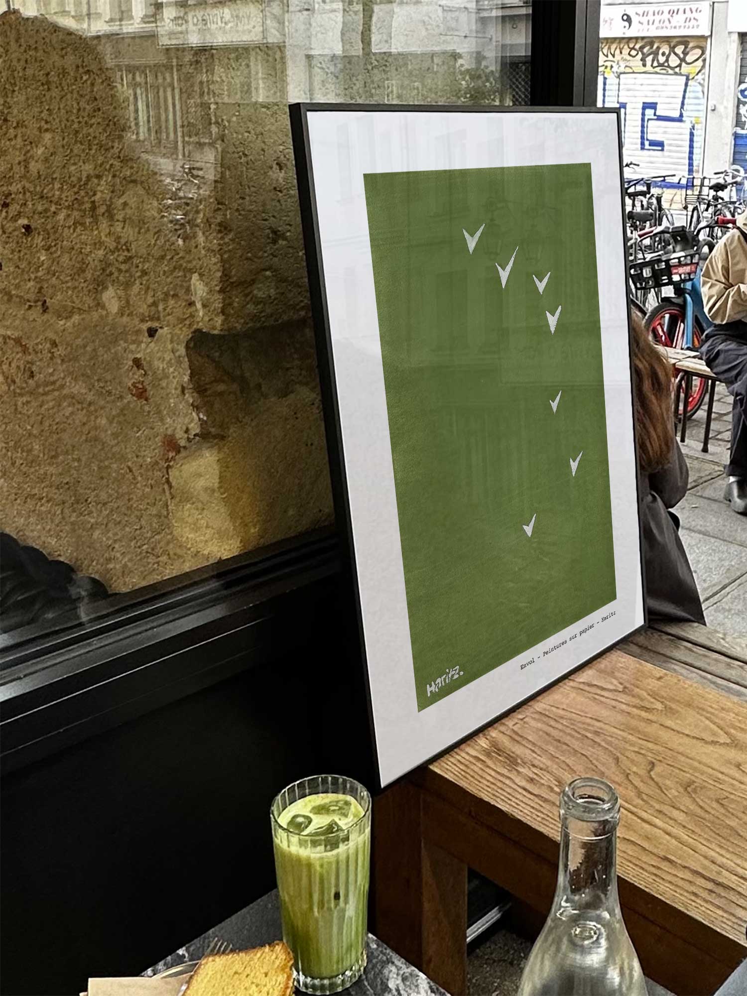 Affiche Envol de Haritz exposée près d'une fenêtre dans un café parisien tendance à côté d'un verre de matcha latte.