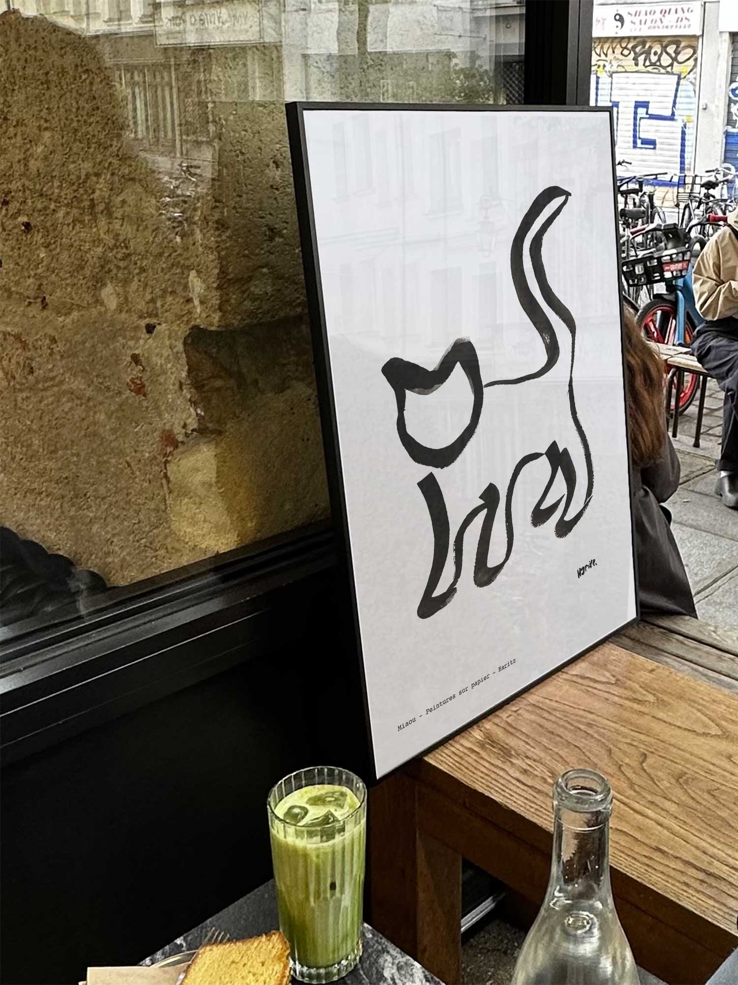 Affiche Miaou de Haritz exposée près d'une fenêtre dans un café parisien tendance à côté d'un verre de matcha latte.