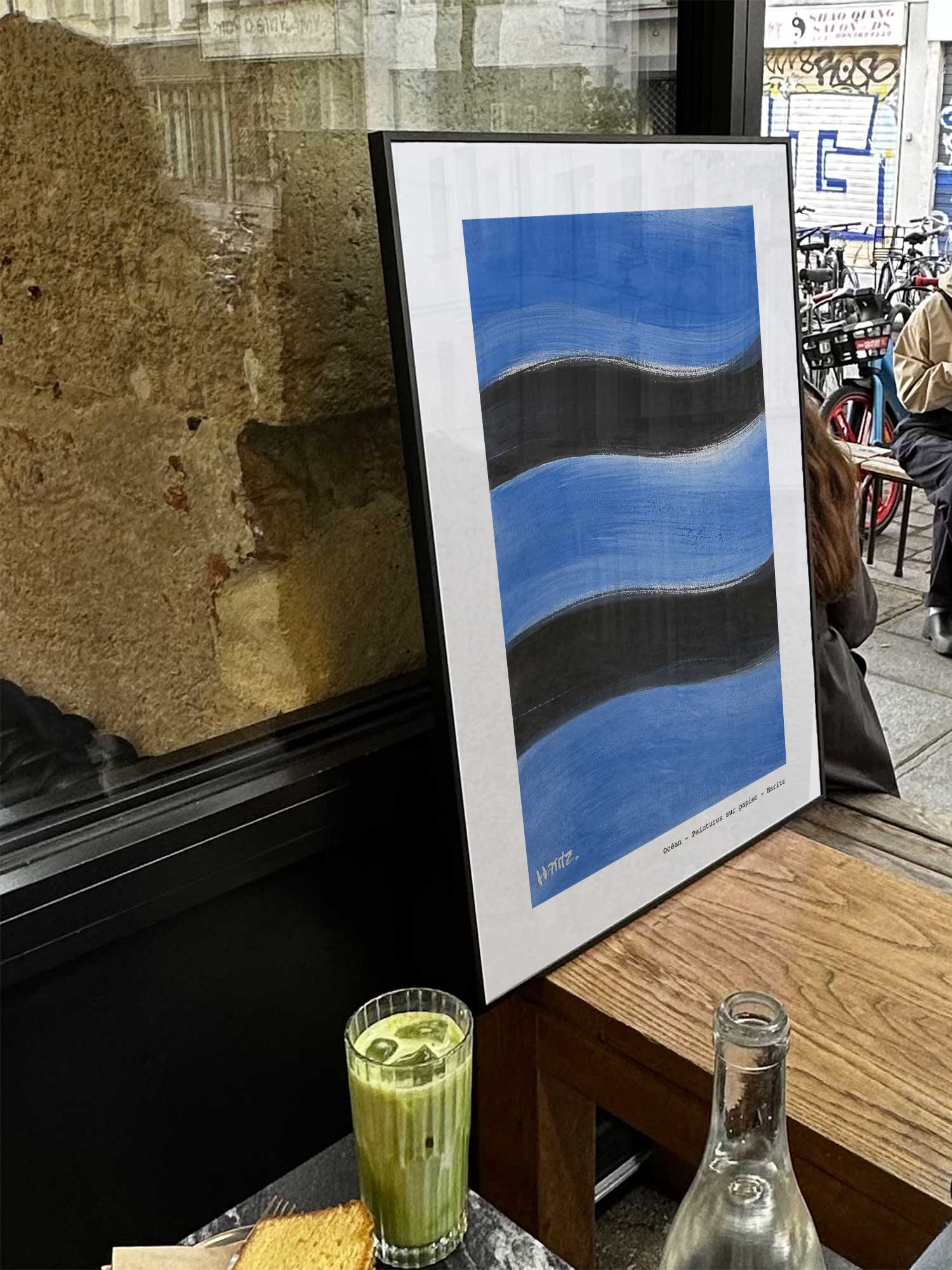Affiche Ocean de Haritz exposée près d'une fenêtre dans un café parisien tendance à côté d'un verre de matcha latte.