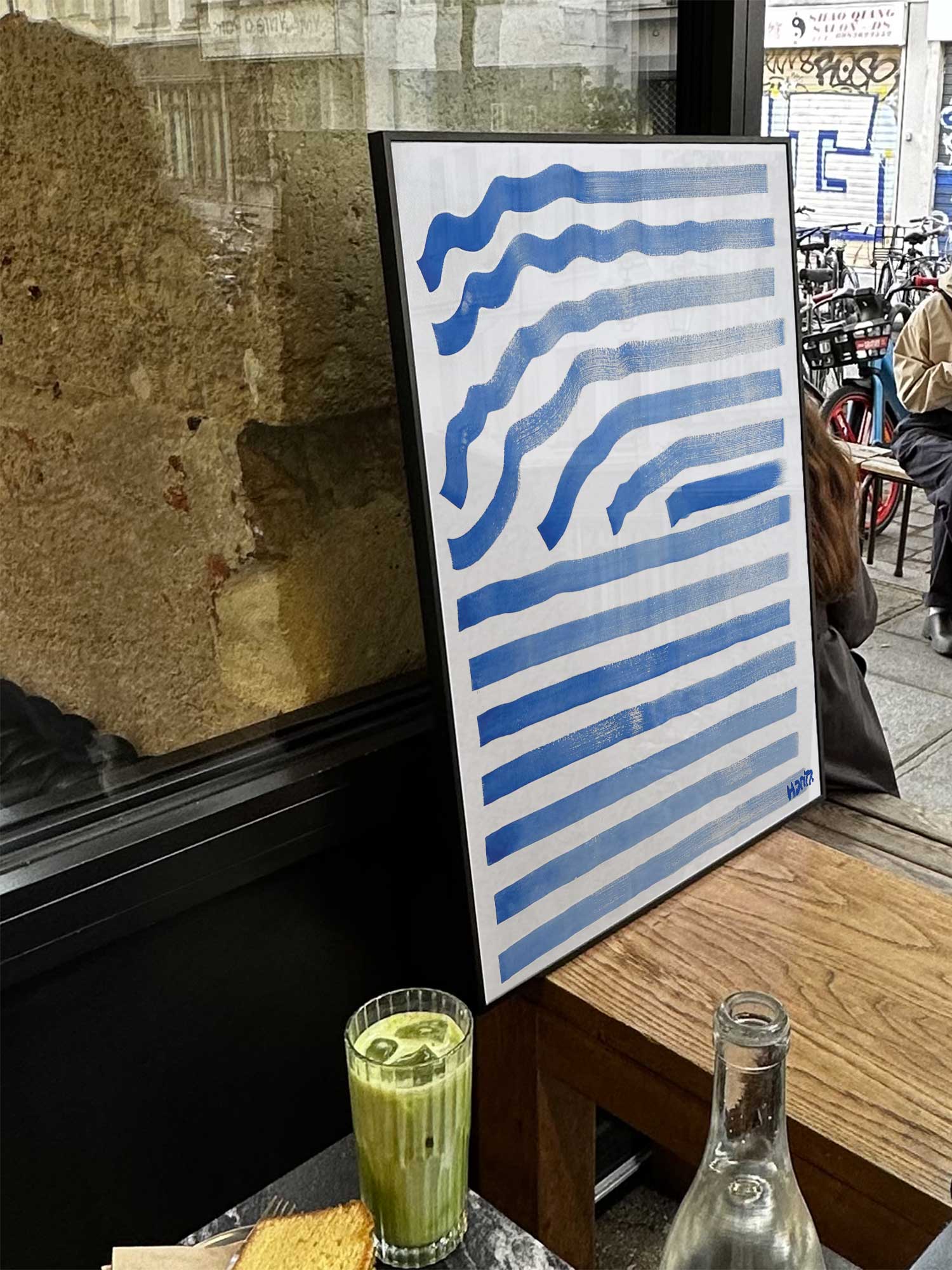 Affiche Ondes bleues de Haritz exposée près d'une fenêtre dans un café parisien tendance à côté d'un verre de matcha latte.