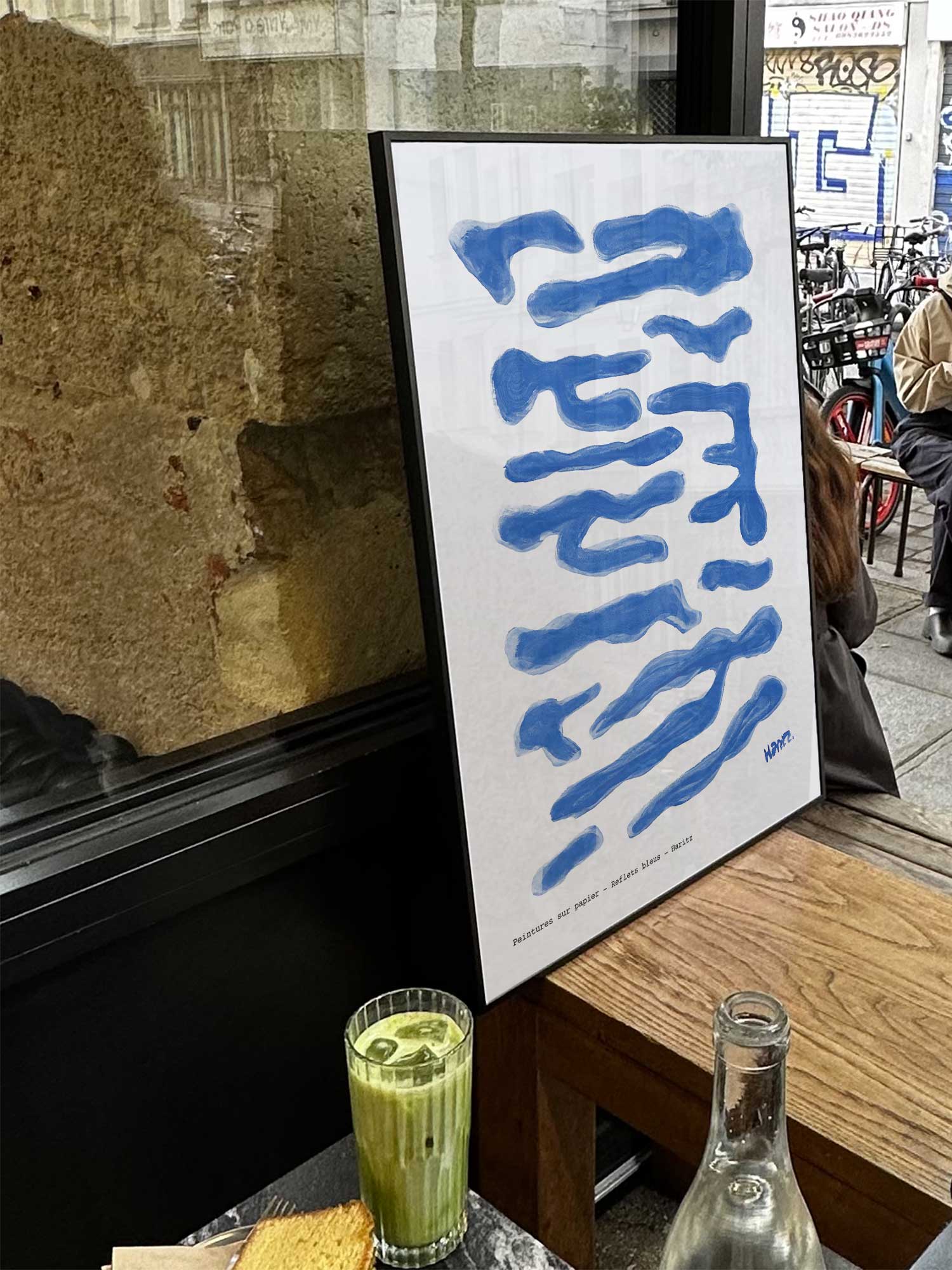 Affiche Reflets bleus de Haritz exposée près d'une fenêtre dans un café parisien tendance à côté d'un verre de matcha latte.