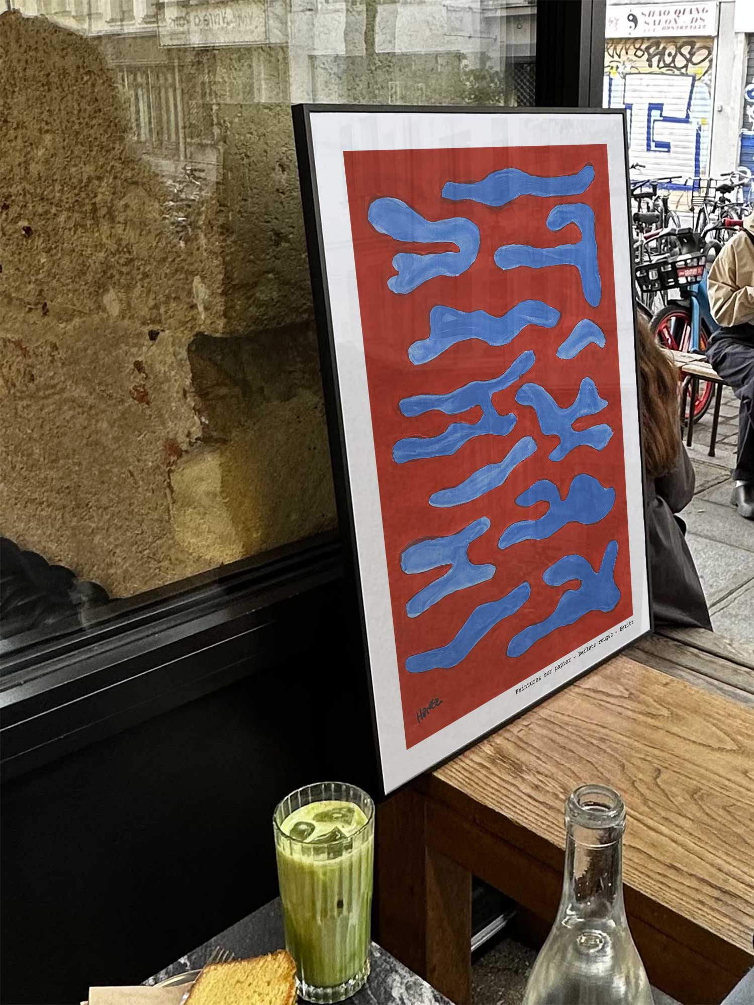 Affiche Reflets rouges de Haritz exposée près d'une fenêtre dans un café parisien tendance à côté d'un verre de matcha latte.