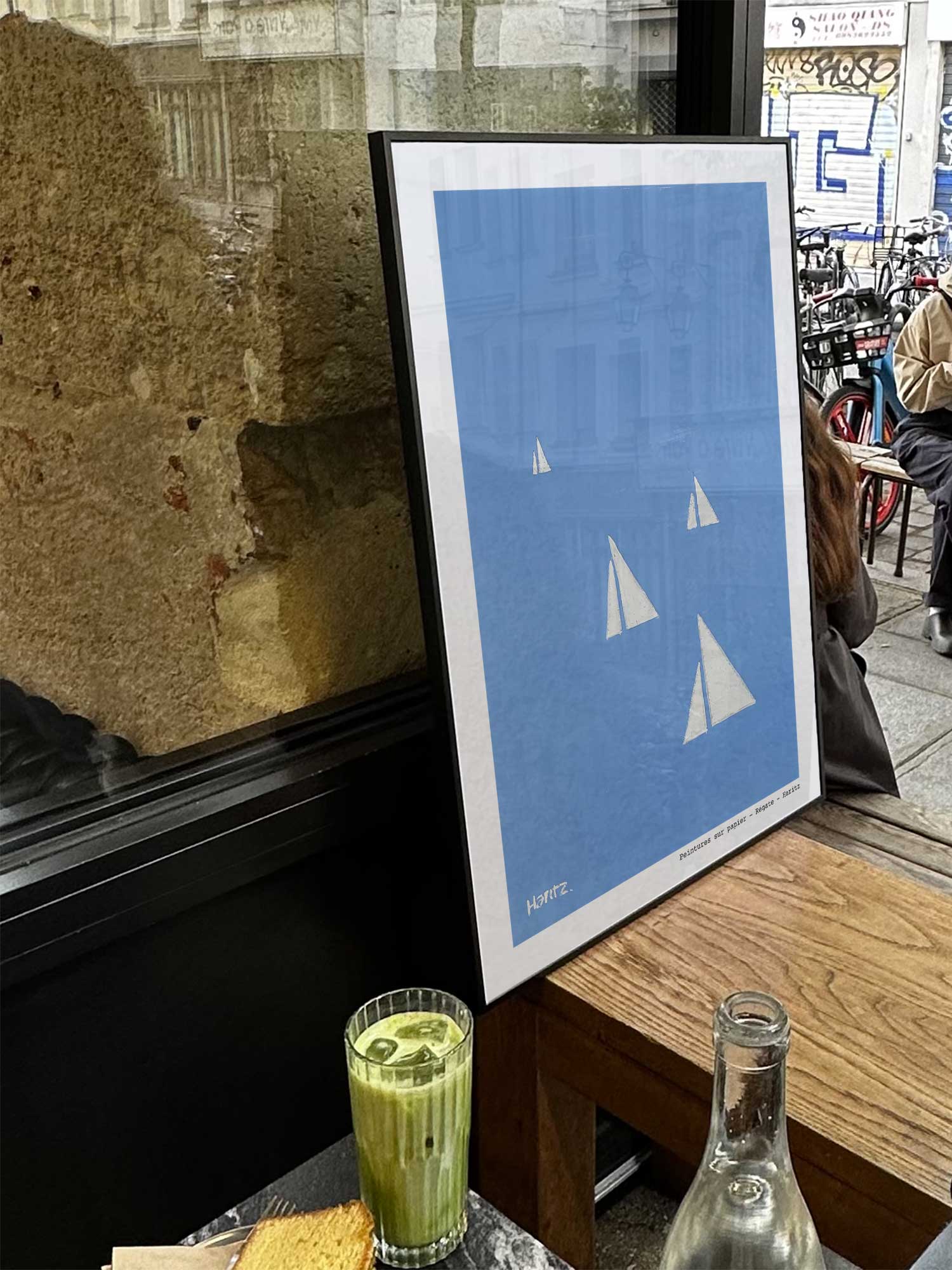 Affiche Régate de Haritz exposée près d'une fenêtre dans un café parisien tendance à côté d'un verre de matcha latte.