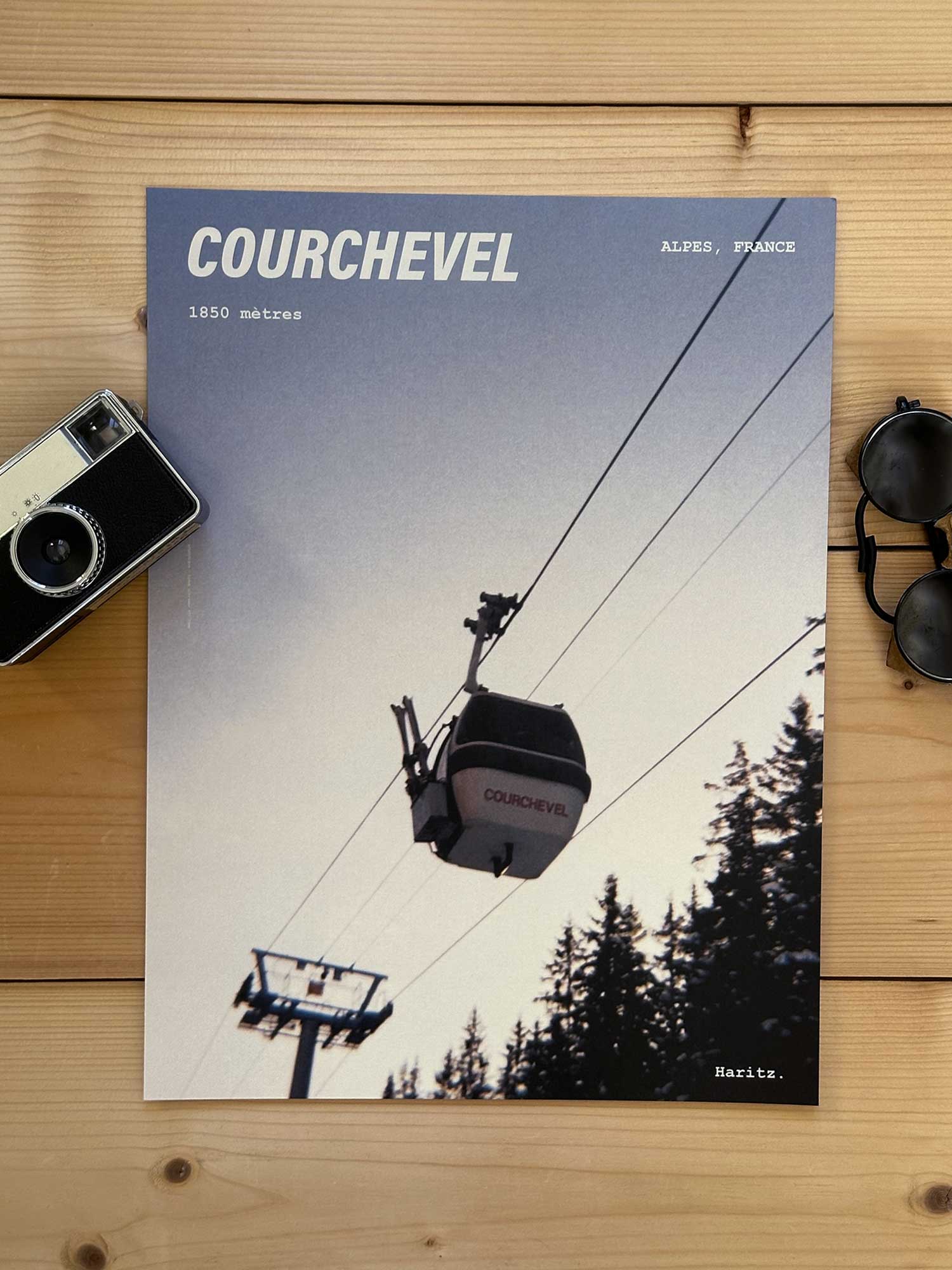 Affiche Courchevel vintage dans un style années 80, sans cadre posée sur parquet bois - À côté d'un ancien appareil photo argentique et d'une paire de lunettes de soleil glacier. Photographie argentique Haritz. L'affiche montre un télécabine de Courchevel