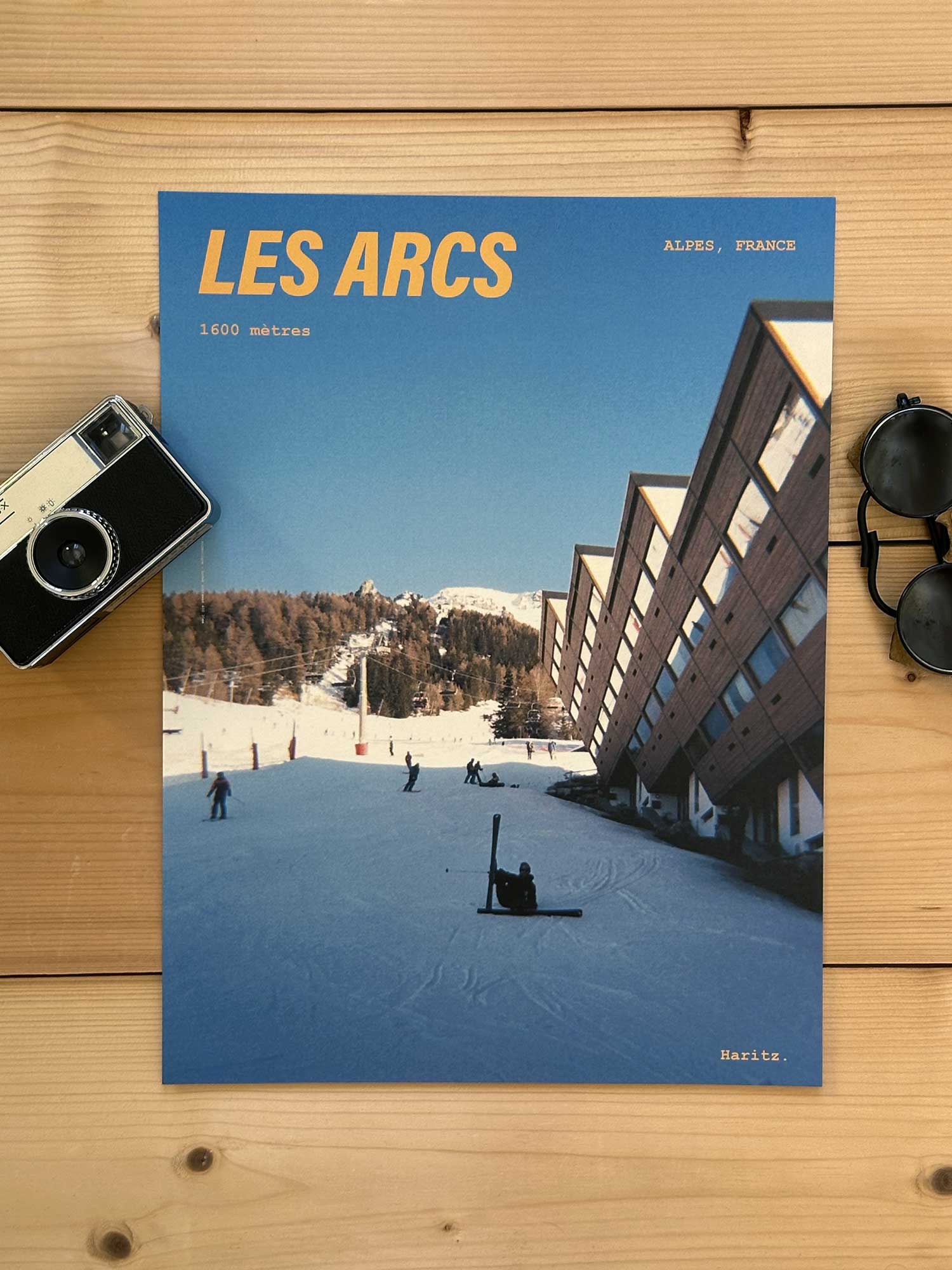 Affiche Les Arcs vintage dans un style années 80, sans cadre posée sur parquet bois - À côté d'un ancien appareil photo argentique et d'une paire de lunettes de soleil glacier. Photographie argentique Haritz. L'affiche montre les Arcs 1600m