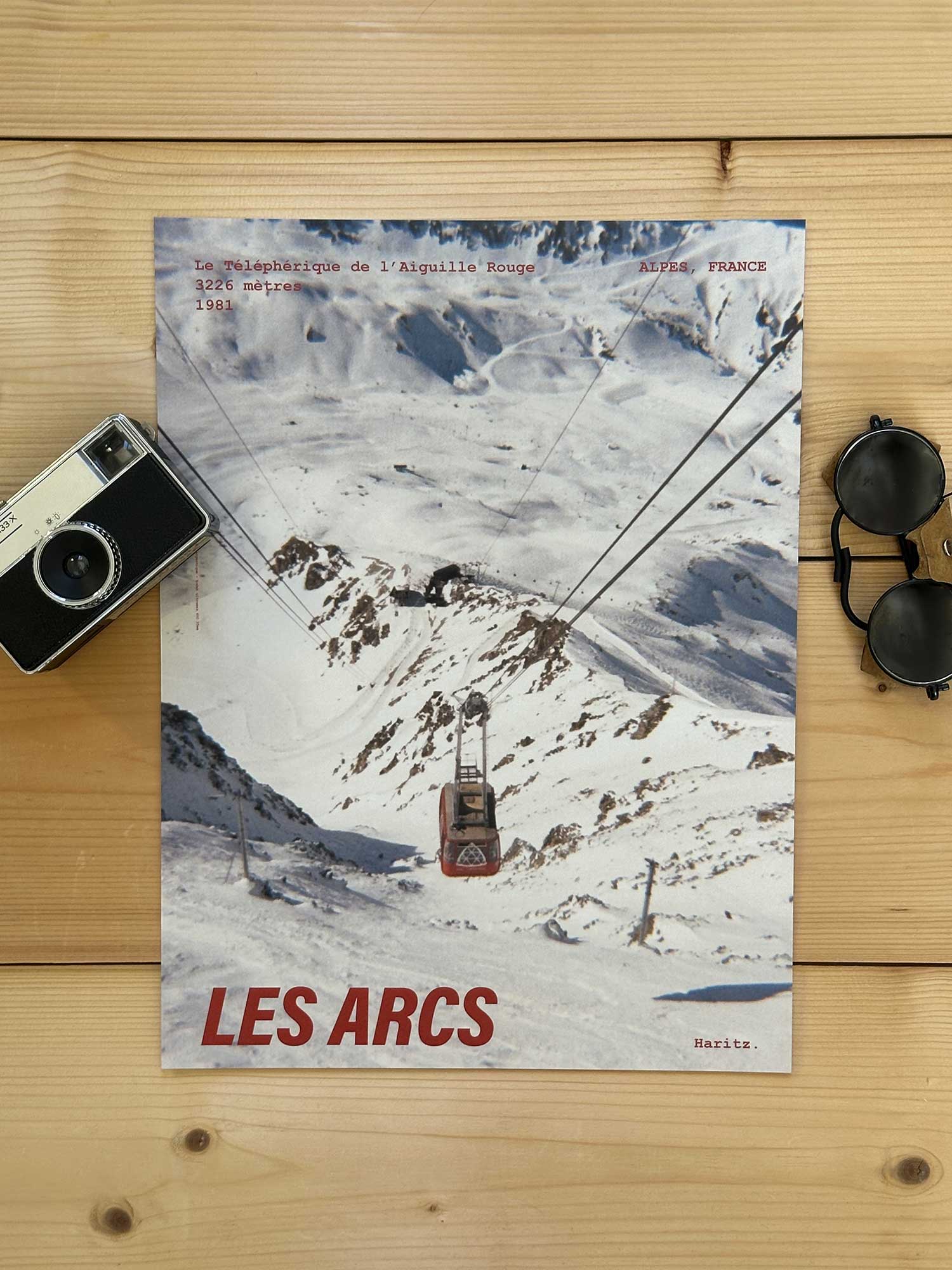Affiche Les Arcs vintage dans un style années 80, sans cadre posée sur parquet bois - À côté d'un ancien appareil photo argentique et d'une paire de lunettes de soleil glacier. Photographie argentique Haritz. L'affiche montre le téléphérique de l'aiguille rouge aux Arcs
