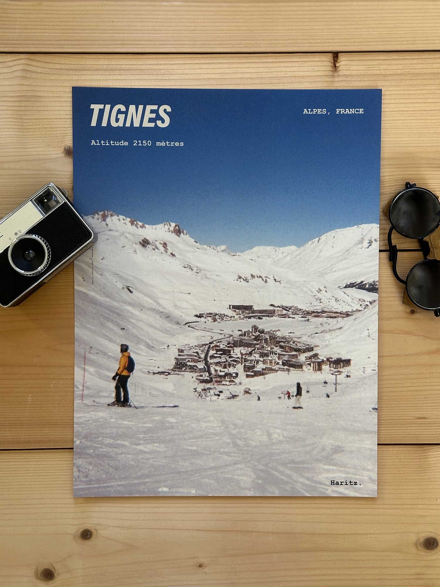 Affiche Tignes vintage dans un style années 80, sans cadre posée sur parquet bois - À côté d'un ancien appareil photo argentique et d'une paire de lunettes de soleil glacier. Photographie argentique Haritz. L'affiche montre le village tignes vu des pistes