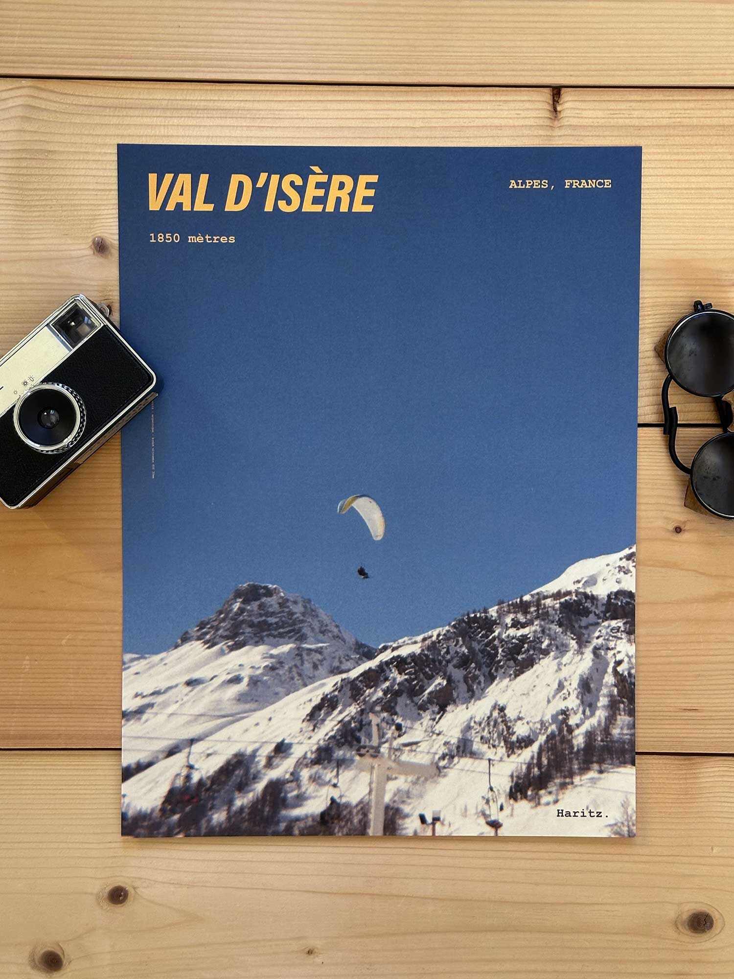 Affiche Val d'Isère vintage dans un style années 80, sans cadre posée sur parquet bois - À côté d'un ancien appareil photo argentique et d'une paire de lunettes de soleil glacier. Photographie argentique Haritz. L'affiche montre un parapente en montagne