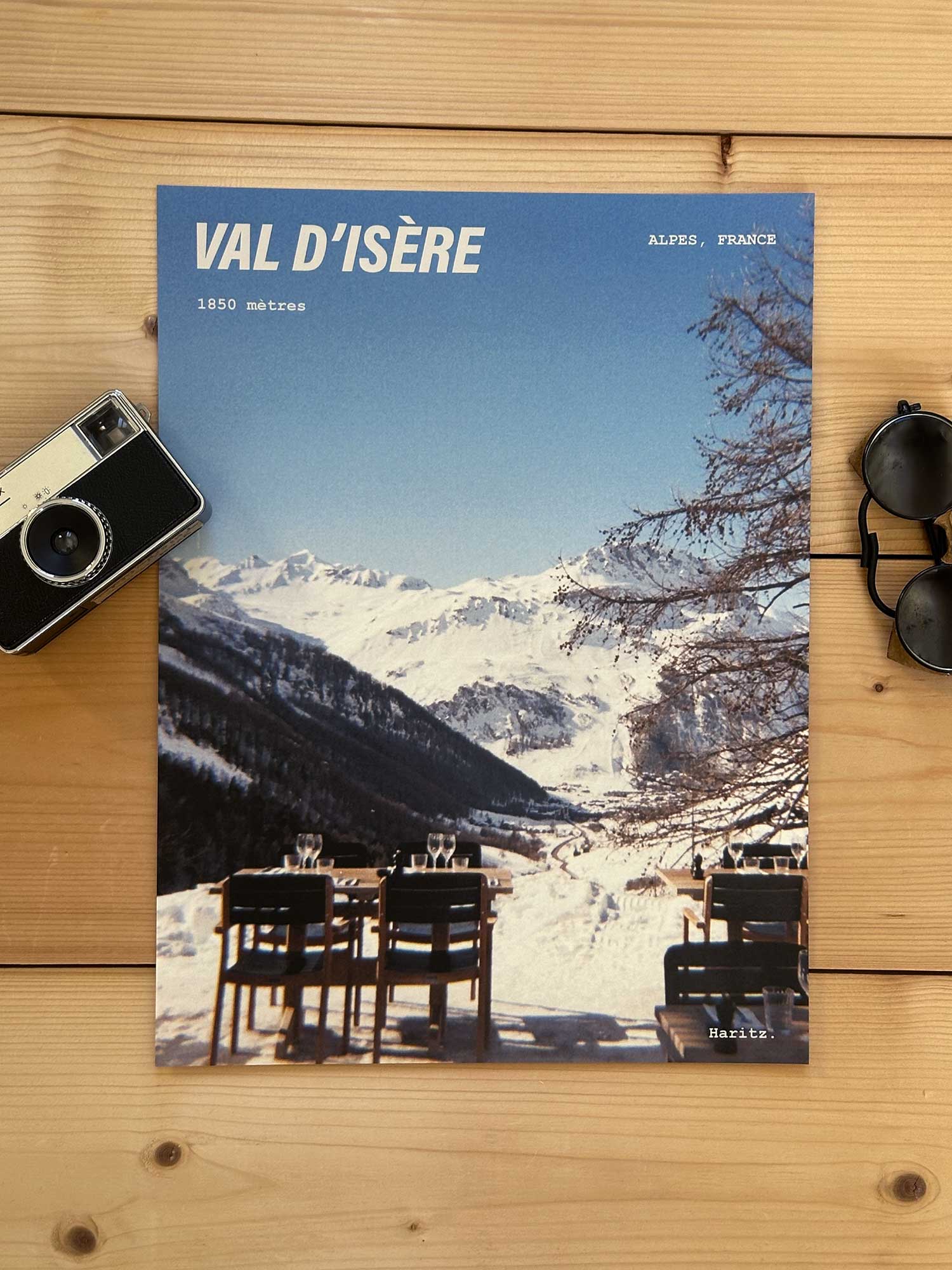 Affiche Val d'Isère vintage dans un style années 80, sans cadre posée sur parquet bois - À côté d'un ancien appareil photo argentique et d'une paire de lunettes de soleil glacier. Photographie argentique Haritz. L'affiche montre une terrasse de restaurant de montagne avec le village de val d'Isère en fond