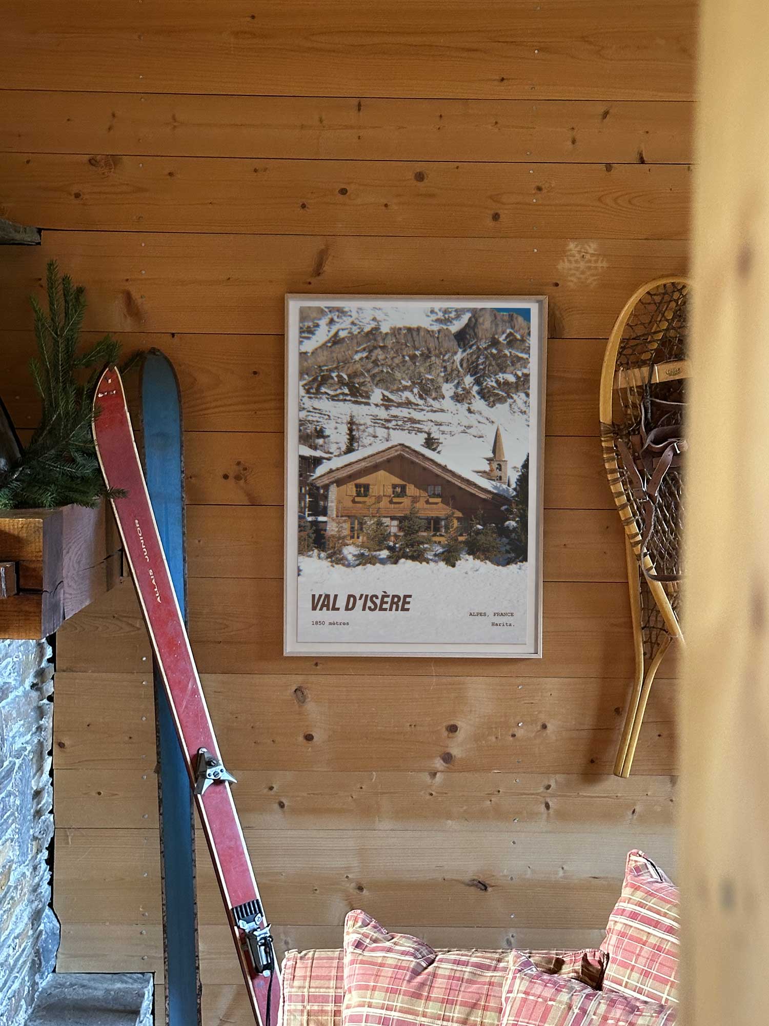 Affiche Val d'Isère vintage encadrée posée sur un mur en bois dans un salon de montagne avec cheminée et canapé — chalet savoyard et clocher du village à 1850 mètres, photographie argentique Haritz
