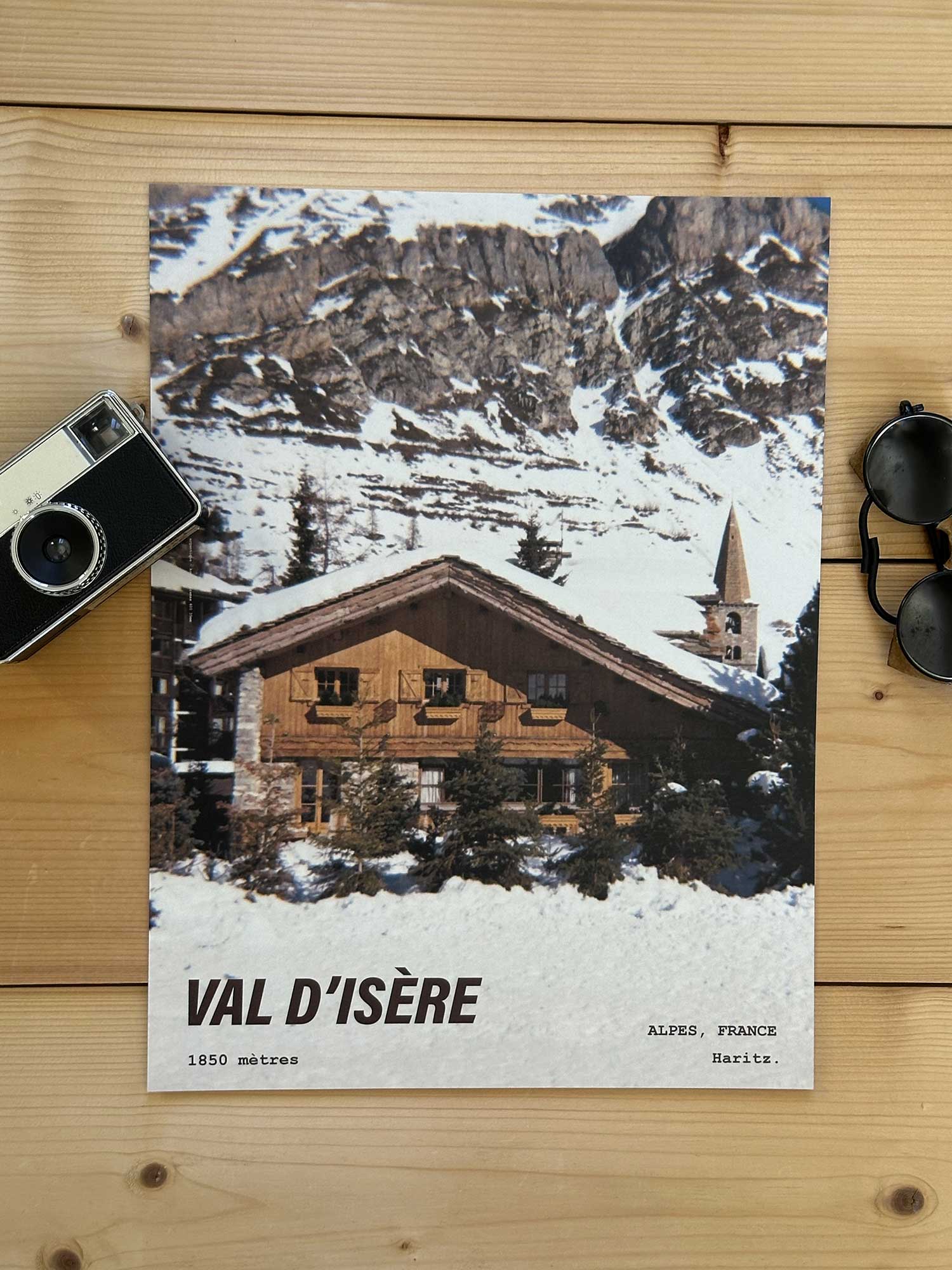 Affiche Val d'Isère vintage dans un style années 80, sans cadre posée sur parquet bois - À côté d'un ancien appareil photo argentique et d'une paire de lunettes de soleil glacier. Photographie argentique Haritz. L'affiche montre le village de Val d'Isère