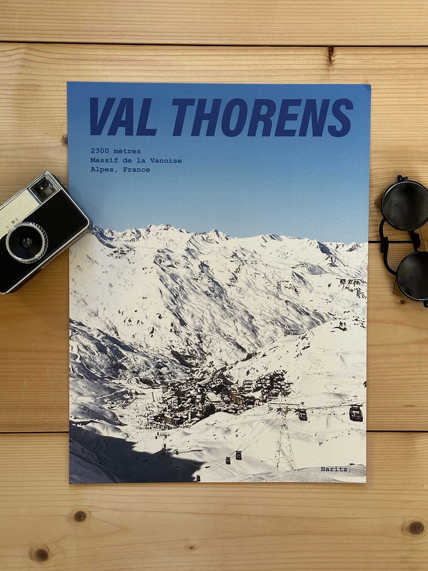 Affiche Val Thorens vintage dans un style années 80, sans cadre posée sur parquet bois - À côté d'un ancien appareil photo argentique et d'une paire de lunettes de soleil glacier. Photographie argentique Haritz. L'affiche montre le village de Val Thorens depuis les pistes
