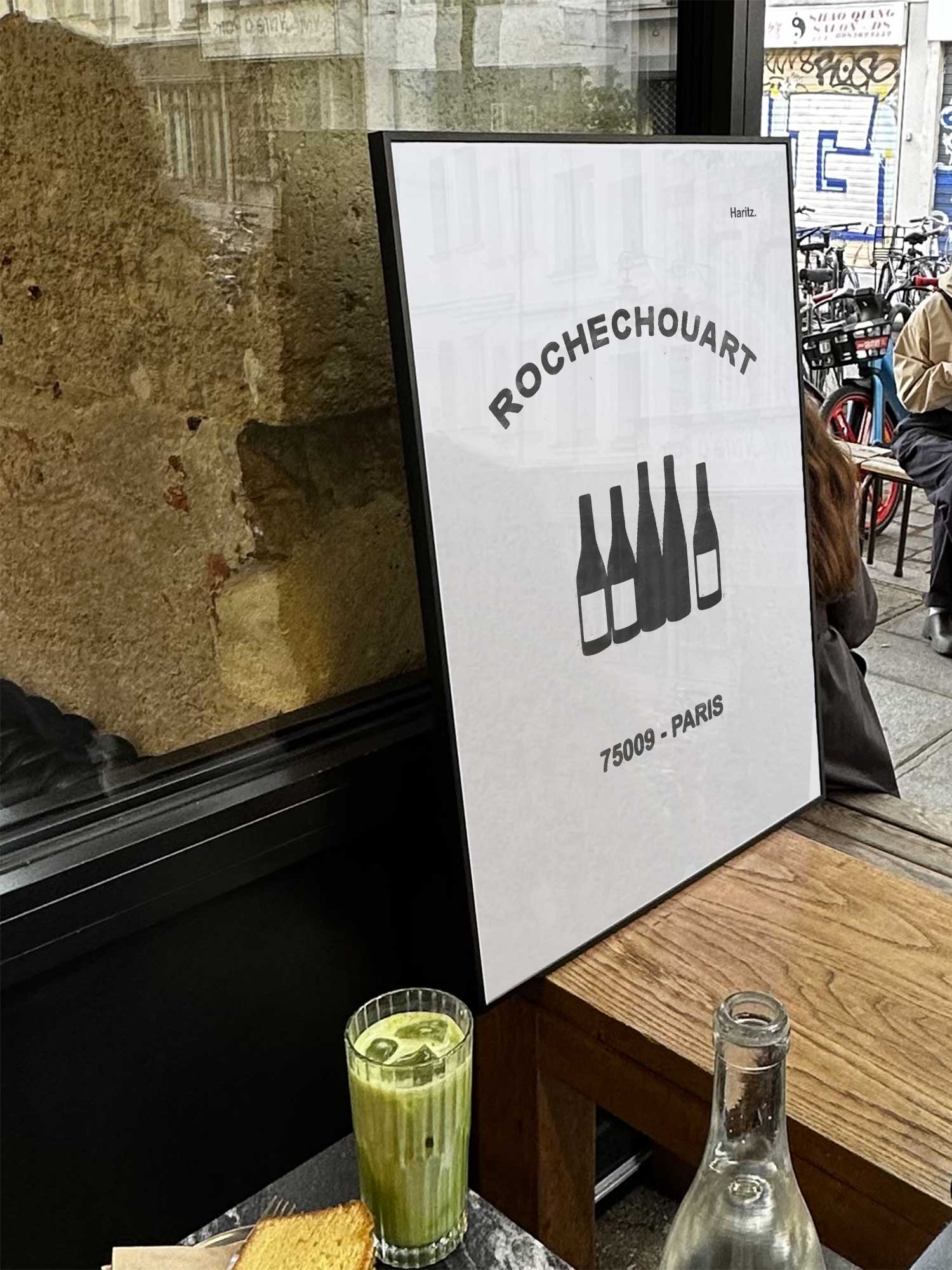Affiche Rochechouart de Haritz exposée près d'une fenêtre dans un café parisien tendance à côté d'un verre de matcha latte.