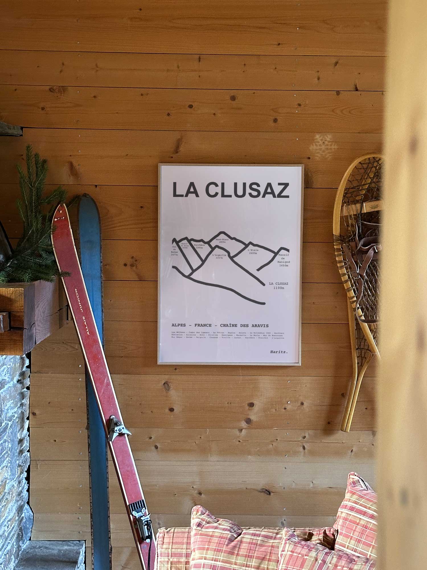 Affiche La Clusaz représentant les sommets de la station, accrochée au mur d’un salon de chalet avec skis vintage et raquettes à neige, décoration montagne Haritz.
