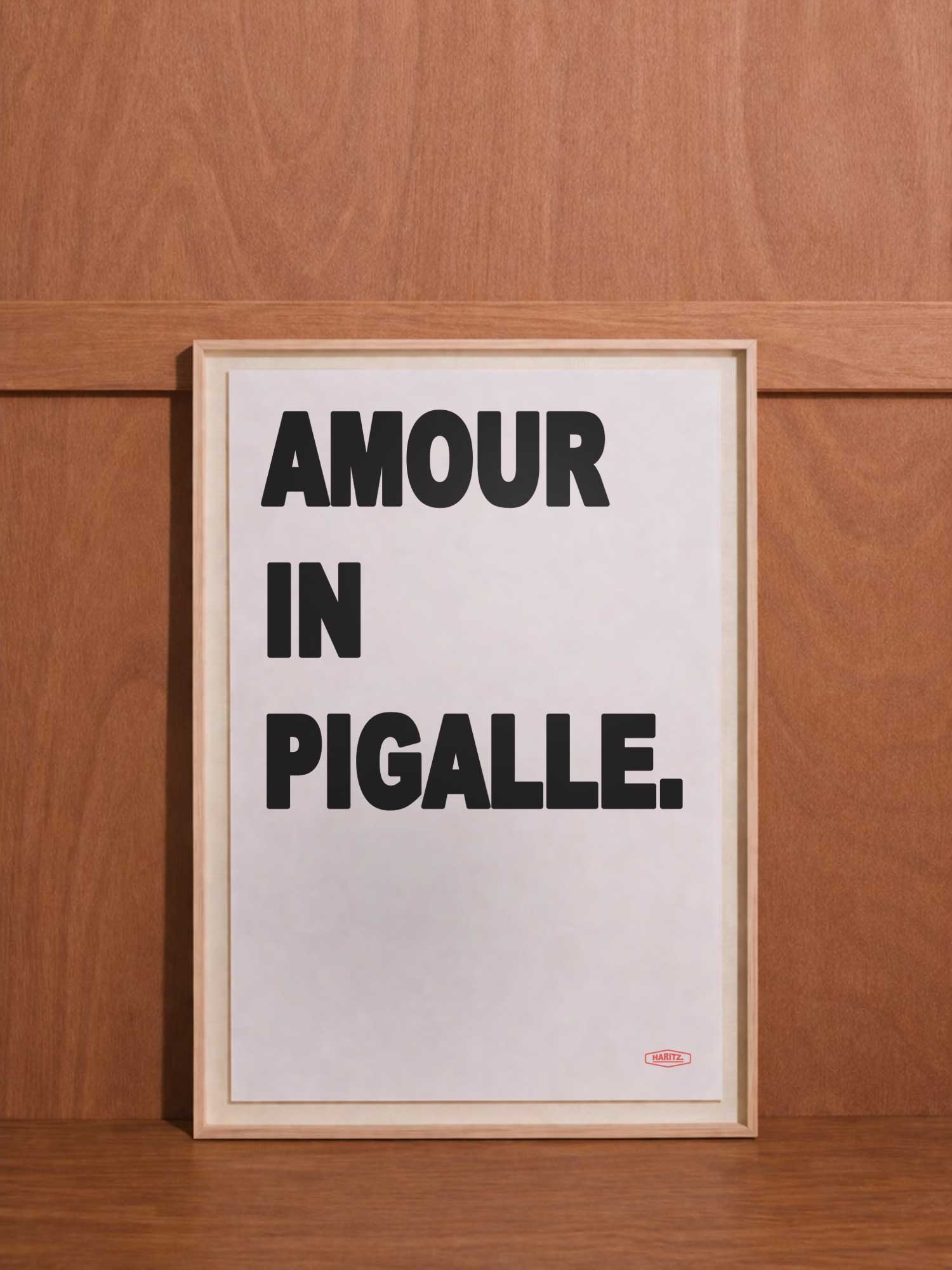 Affiche contemporaine de décoration Haritz avec le texte "AMOUR IN PIGALLE." en lettres noires grasses sur fond blanc, encadrée et posée contre un mur en bois