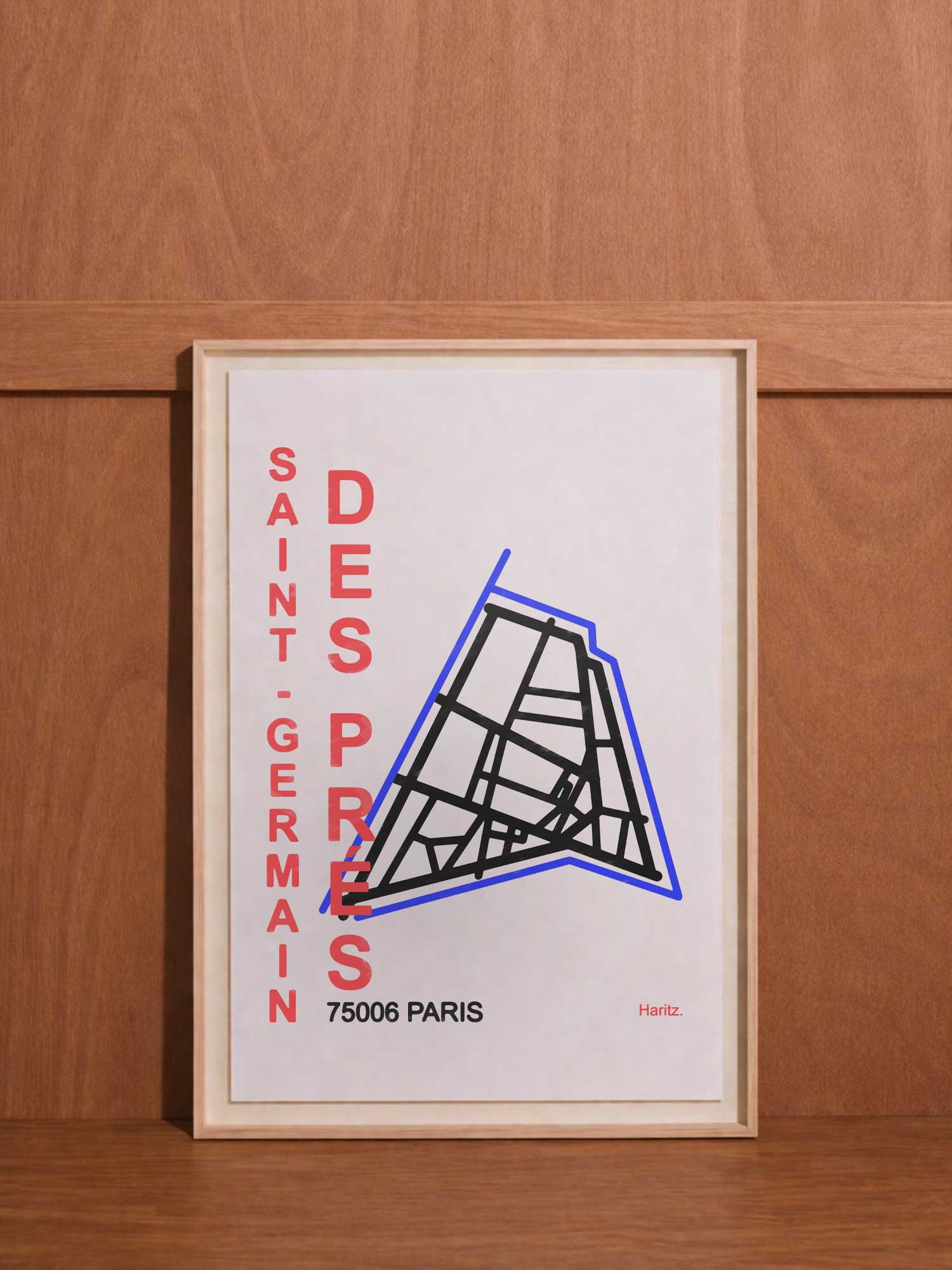 Affiche contemporaine de décoration Haritz de Saint-Germain-des-Prés avec plan de quartier stylisé en noir et bleu, texte vertical rouge "SAINT-GERMAIN DES PRÉS" et "75006 PARIS", encadrée et posée contre un mur en bois