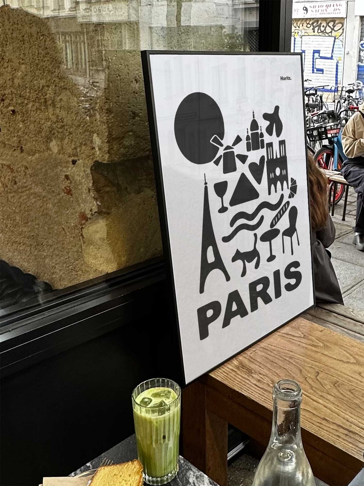 Affiche Paris, version noire et blanche de Haritz exposée près d'une fenêtre dans un café parisien tendance à côté d'un verre de matcha latte.