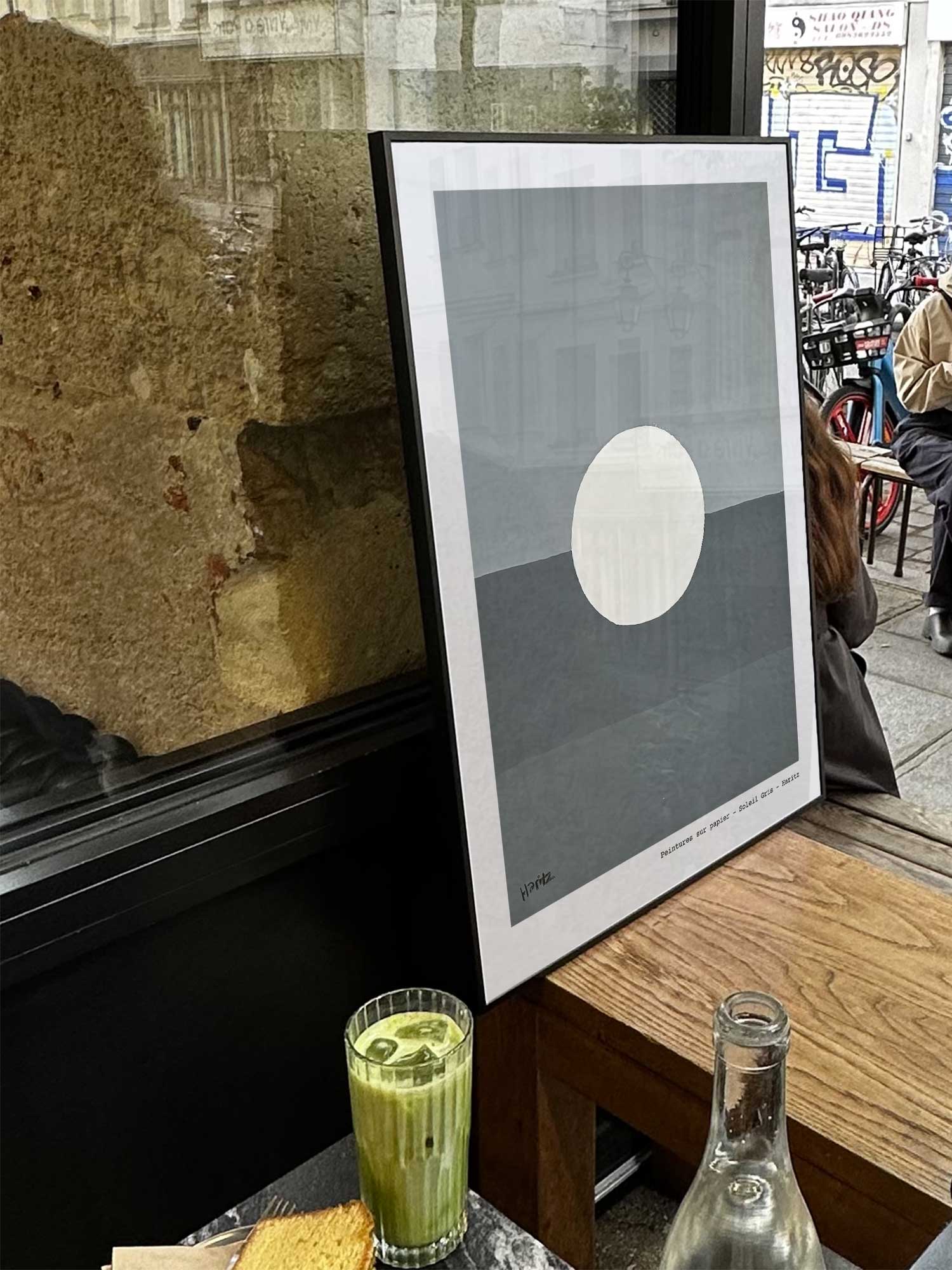 Affiche Lune de Haritz exposée près d'une fenêtre dans un café parisien tendance à côté d'un verre de matcha latte.