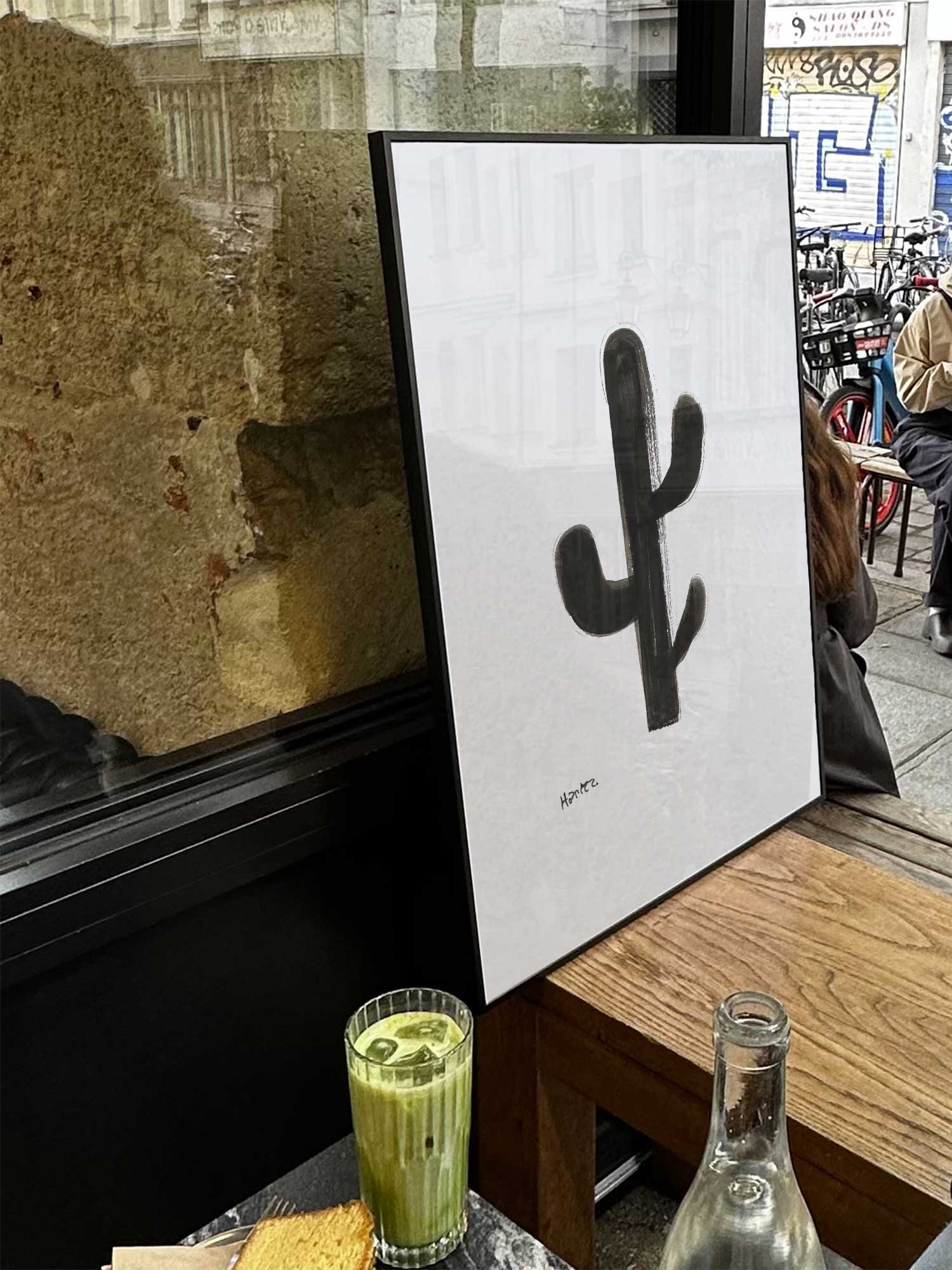Affiche Cactus de Haritz exposée près d'une fenêtre dans un café parisien tendance à côté d'un verre de matcha latte.
