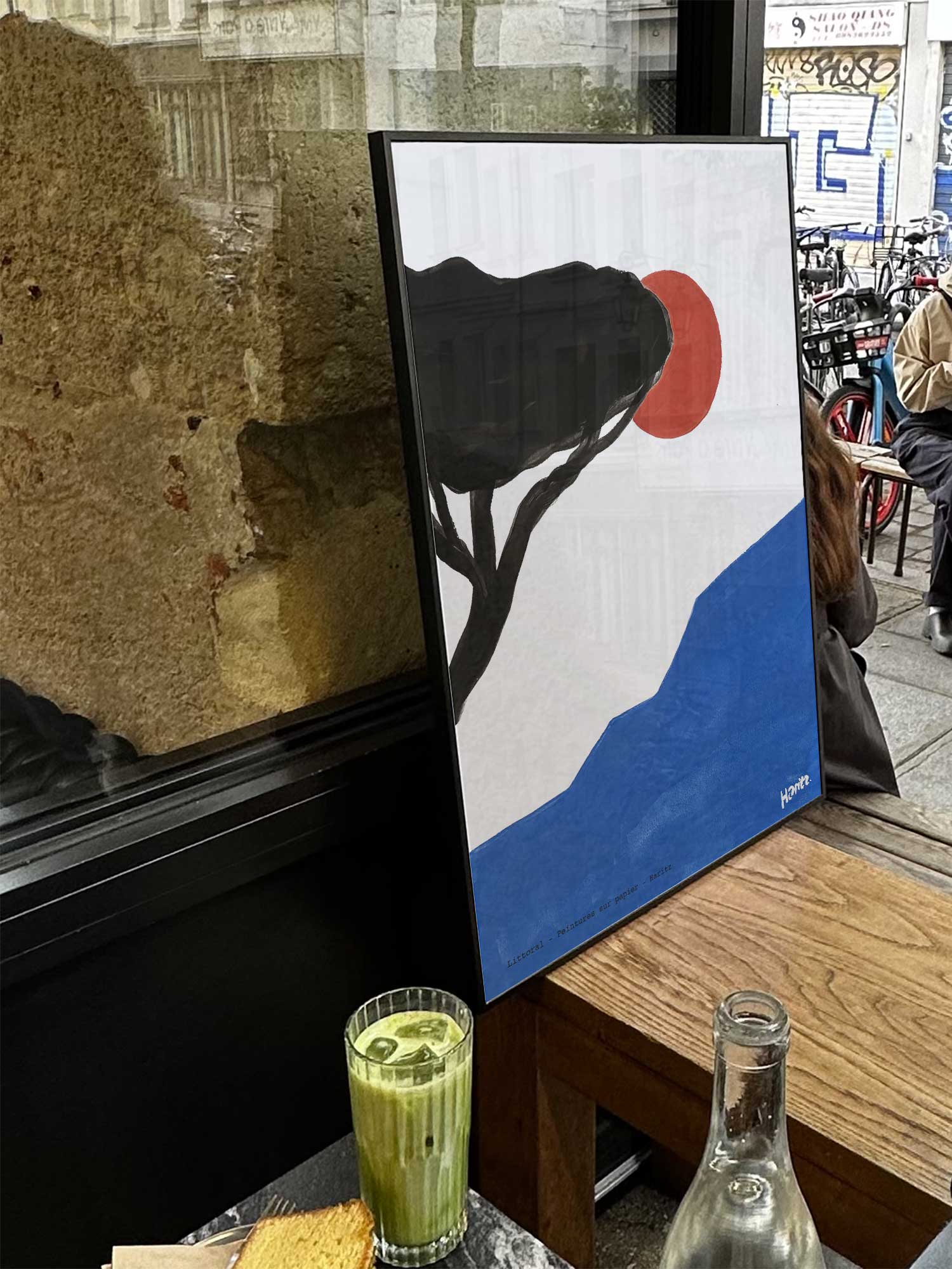 Affiche Littoral de Haritz exposée près d'une fenêtre dans un café parisien tendance à côté d'un verre de matcha latte.