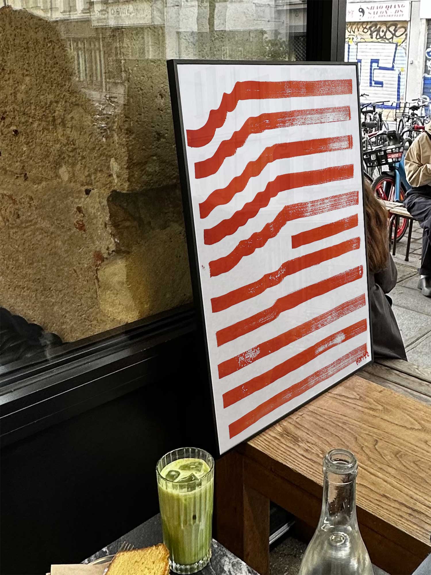 Affiche Ondes rouges de Haritz exposée près d'une fenêtre dans un café parisien tendance à côté d'un verre de matcha latte.