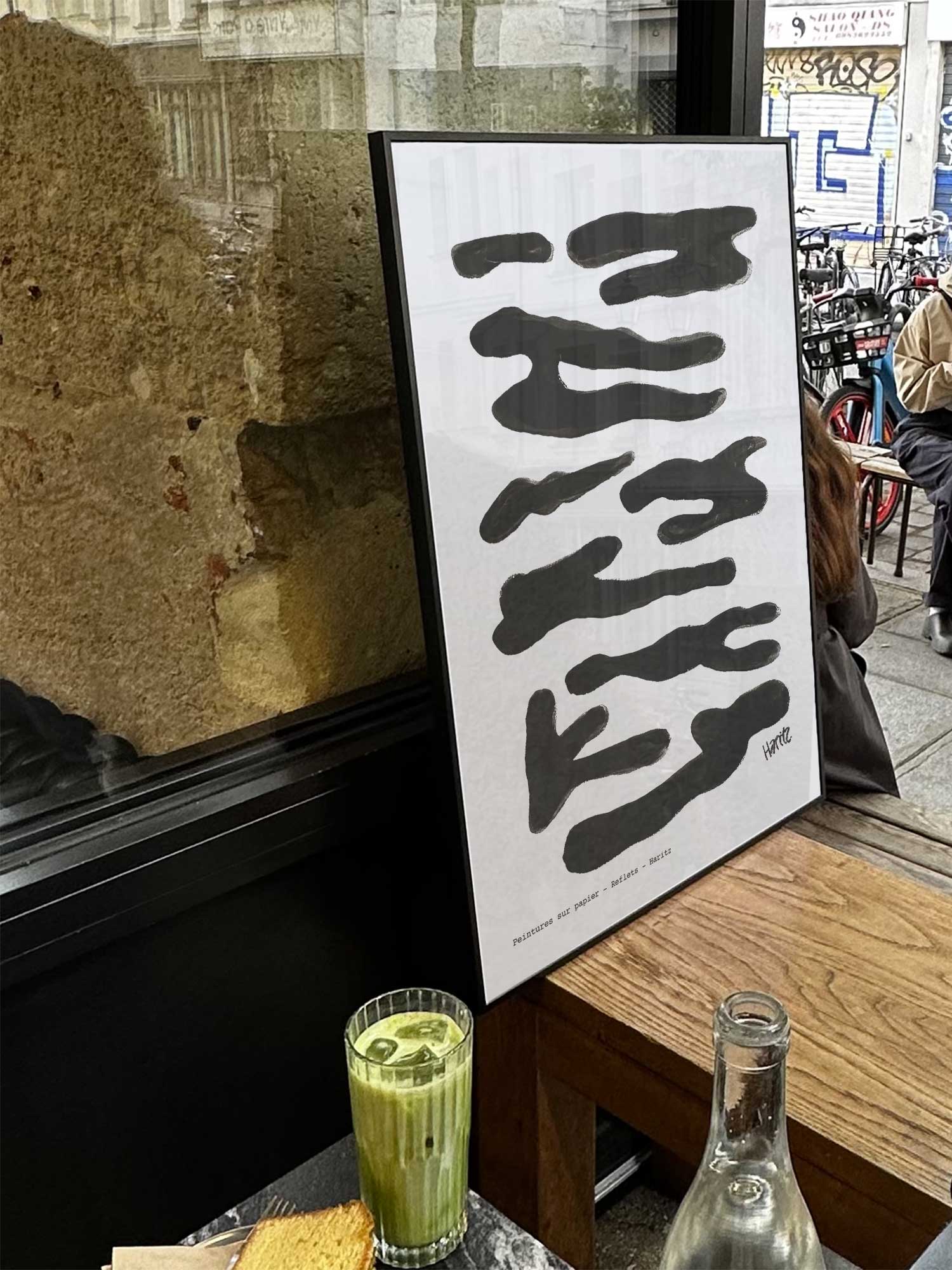 Affiche Reflets noirs de Haritz exposée près d'une fenêtre dans un café parisien tendance à côté d'un verre de matcha latte.