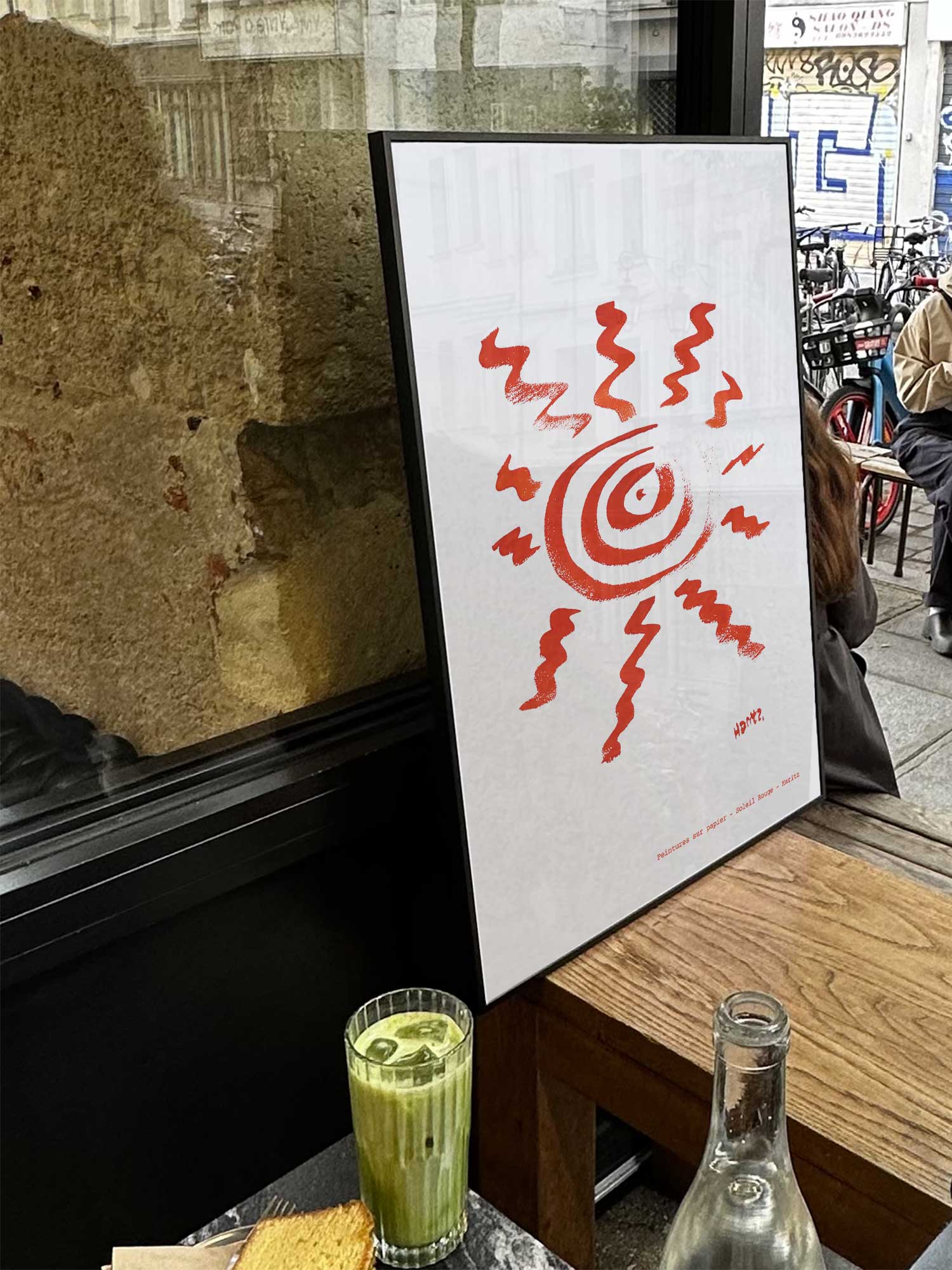 Affiche Soleil rouge de Haritz exposée près d'une fenêtre dans un café parisien tendance à côté d'un verre de matcha latte.