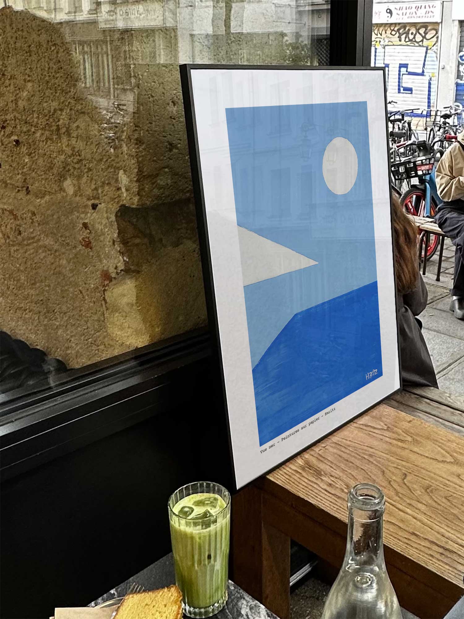 Affiche Vue mer de Haritz exposée près d'une fenêtre dans un café parisien tendance à côté d'un verre de matcha latte.