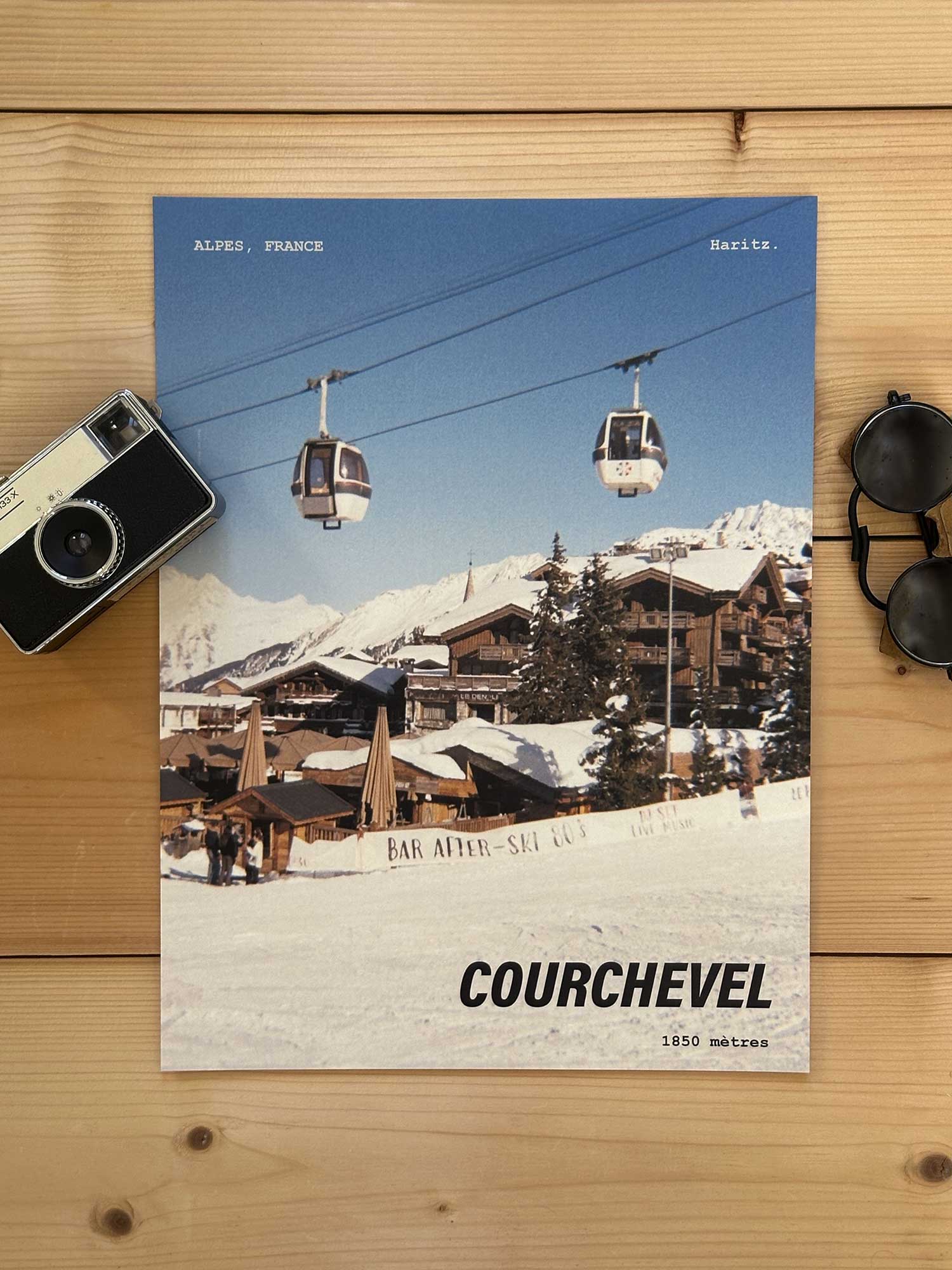 Affiche Courchevel vintage dans un style années 80, sans cadre posée sur parquet bois - À côté d'un ancien appareil photo argentique et d'une paire de lunettes de soleil glacier. Photographie argentique Haritz. L'affiche montre le village Courchevel 1850 avec deux télécabines