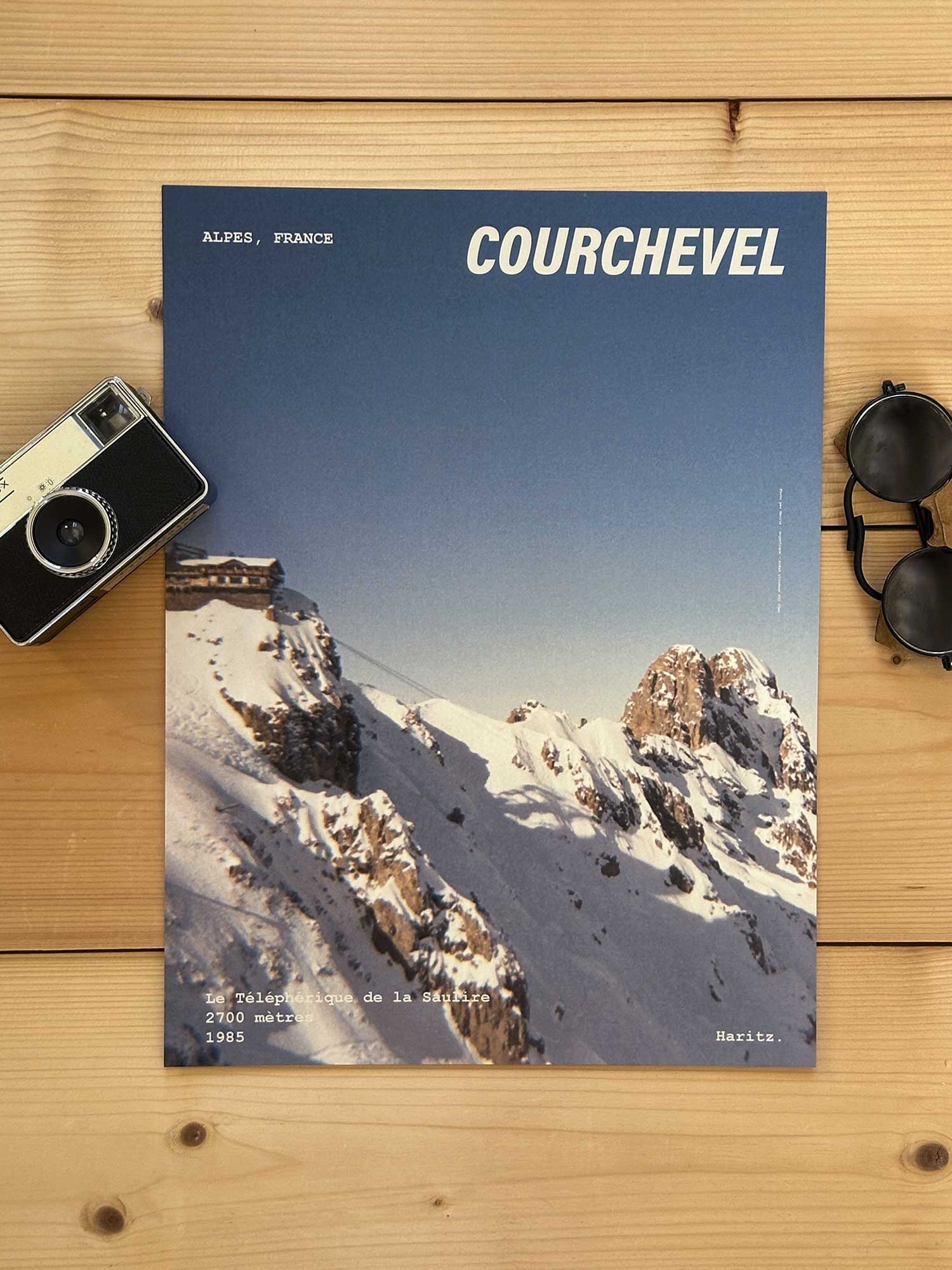 Affiche Courchevel vintage dans un style années 80, sans cadre posée sur parquet bois - À côté d'un ancien appareil photo argentique et d'une paire de lunettes de soleil glacier. Photographie argentique Haritz. L'affiche montre le téléphérique de la Saulire à courchevel