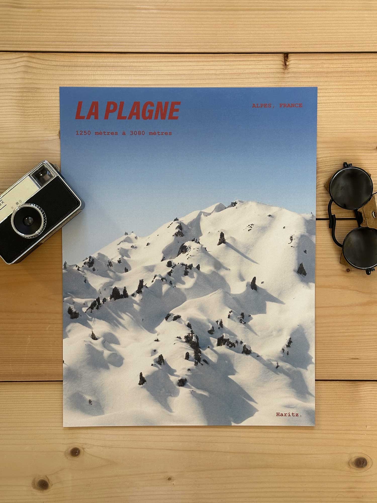 Affiche La Plagne vintage dans un style années 80, sans cadre posée sur parquet bois - À côté d'un ancien appareil photo argentique et d'une paire de lunettes de soleil glacier. Photographie argentique Haritz. L'affiche montre une montagne enneigée de La Plagne 