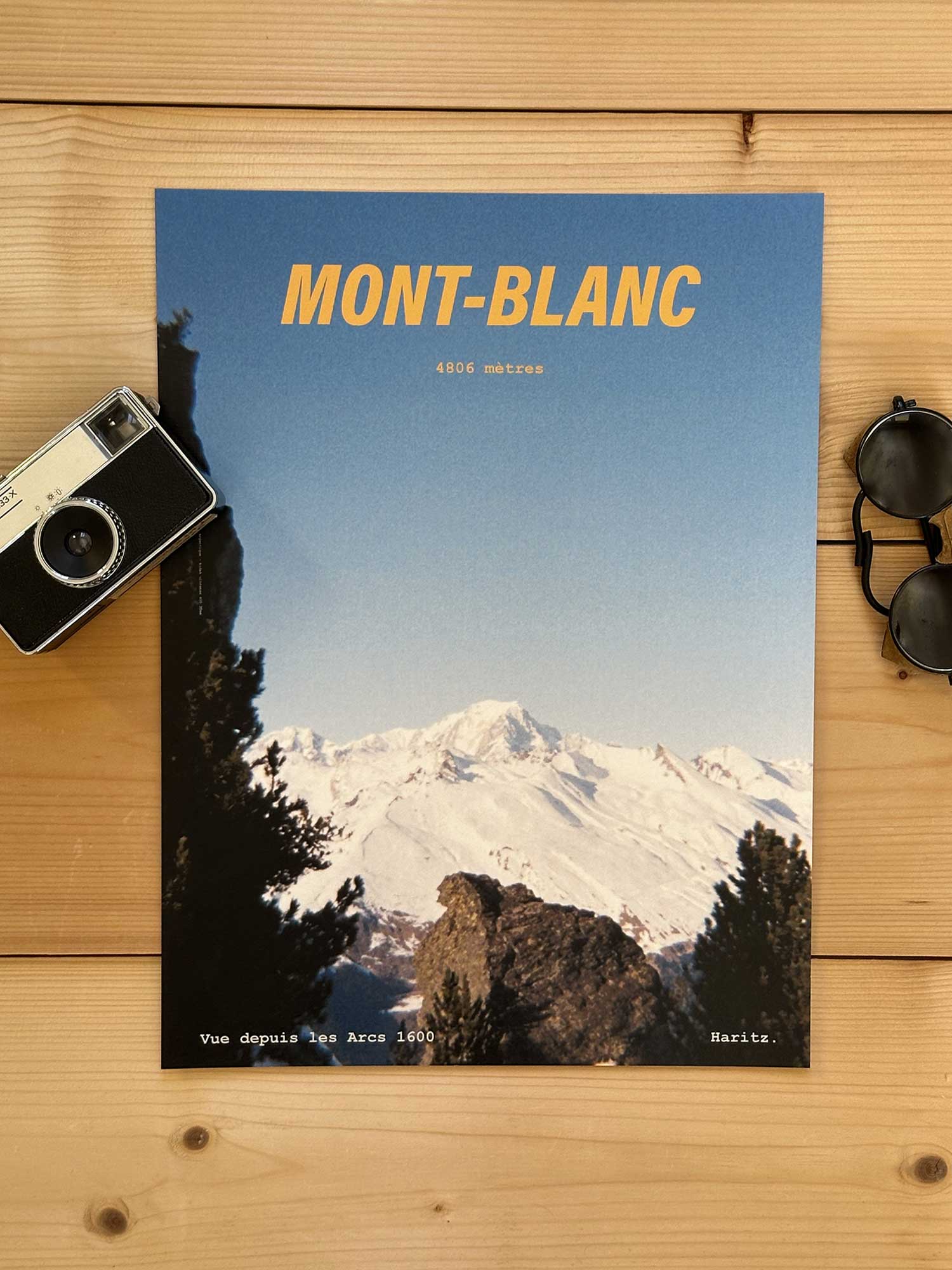 Affiche Mont-Blanc vintage dans un style années 80, sans cadre posée sur parquet bois - À côté d'un ancien appareil photo argentique et d'une paire de lunettes de soleil glacier. Photographie argentique Haritz. L'affiche montre le Mont Blanc vu de loin