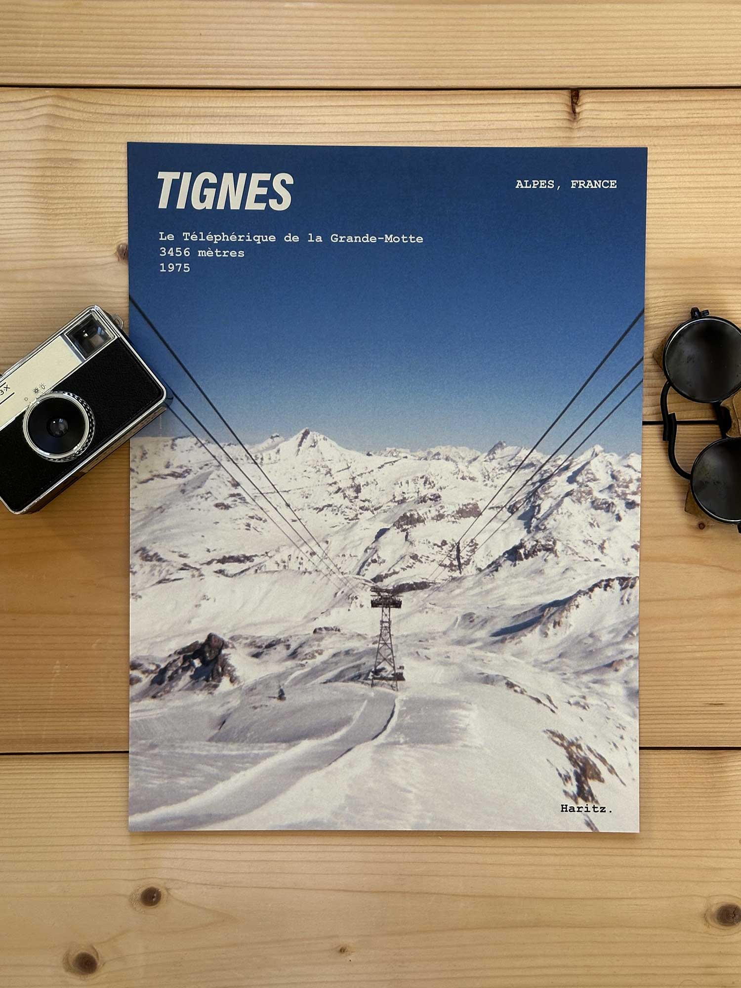 Affiche Tignes vintage dans un style années 80, sans cadre posée sur parquet bois - À côté d'un ancien appareil photo argentique et d'une paire de lunettes de soleil glacier. Photographie argentique Haritz. L'affiche montre le téléphérique de la grande motte à tignes