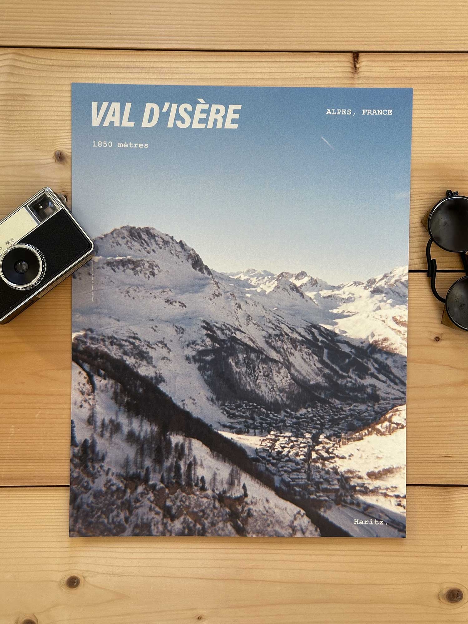 Affiche Val d'Isère vintage dans un style années 80, sans cadre posée sur parquet bois - À côté d'un ancien appareil photo argentique et d'une paire de lunettes de soleil glacier. Photographie argentique Haritz. L'affiche montre le village de Val d'Isère vu du haut