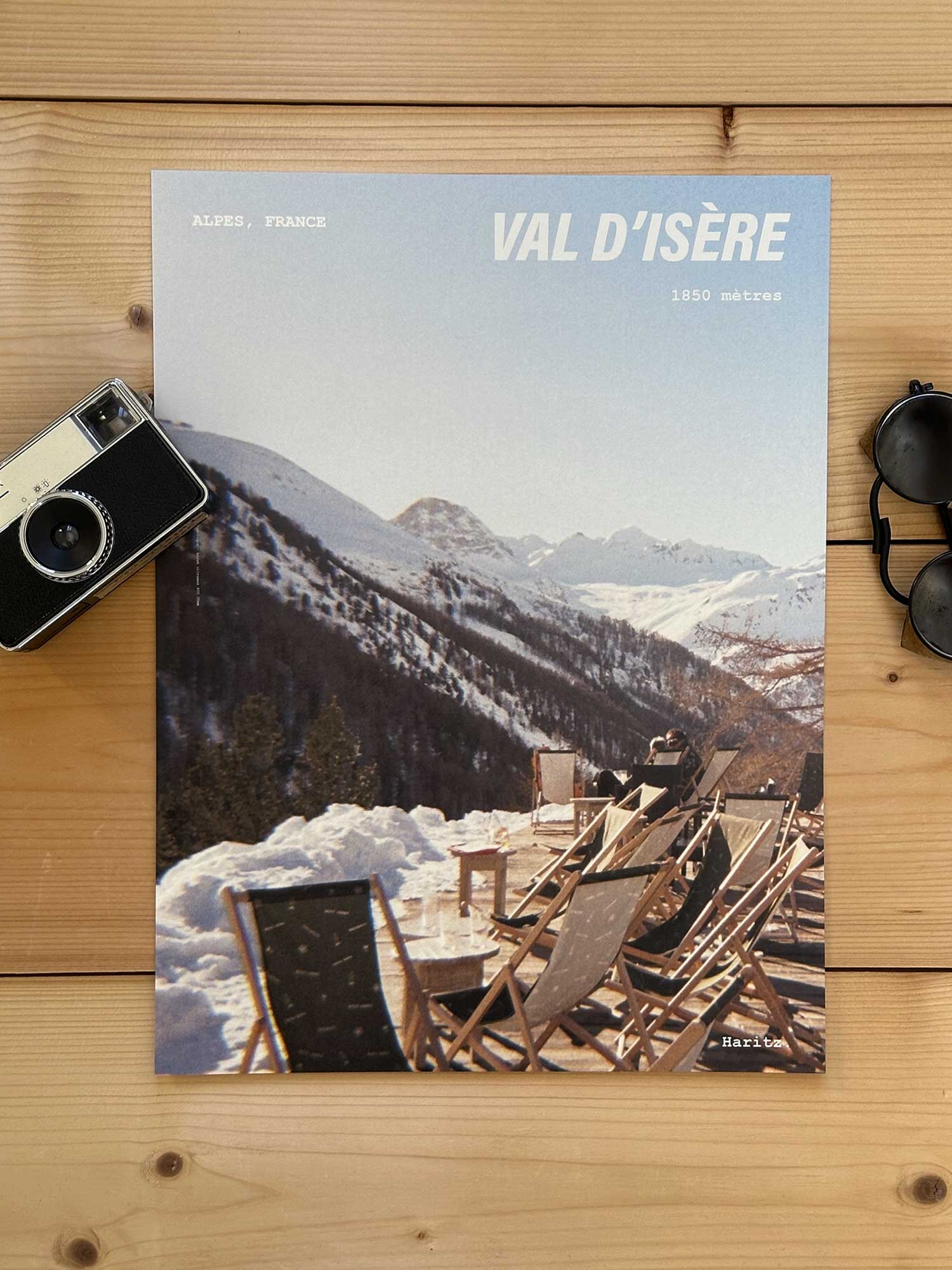 Affiche Val d'Isère vintage dans un style années 80, sans cadre posée sur parquet bois - À côté d'un ancien appareil photo argentique et d'une paire de lunettes de soleil glacier. Photographie argentique Haritz. L'affiche montre une terrasse ensoleillée de montagne à Val d'Isère