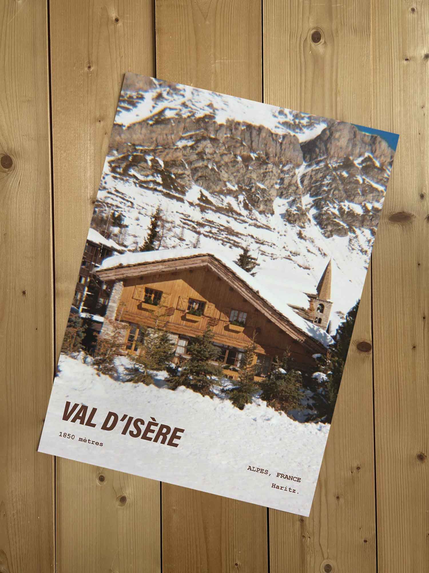 Affiche Val d'Isère vintage sans cadre posée sur parquet bois — chalet et clocher du vieux village sous la neige, photographie argentique Haritz