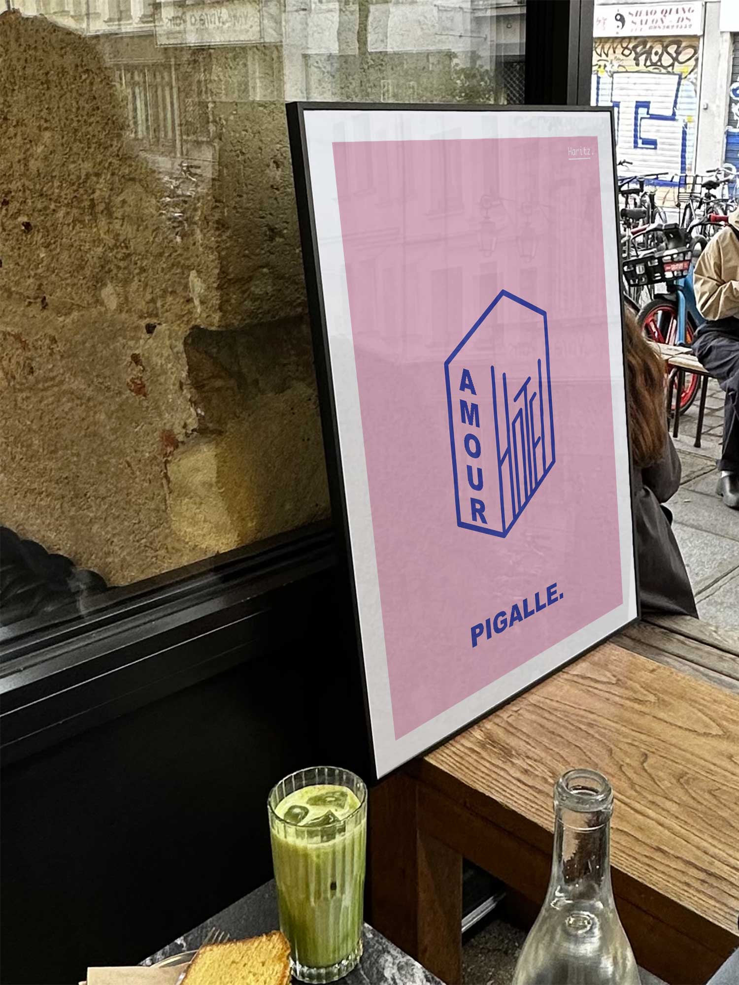 Affiche Hotel Amour de Haritz exposée près d'une fenêtre dans un café parisien tendance à côté d'un verre de matcha latte.
