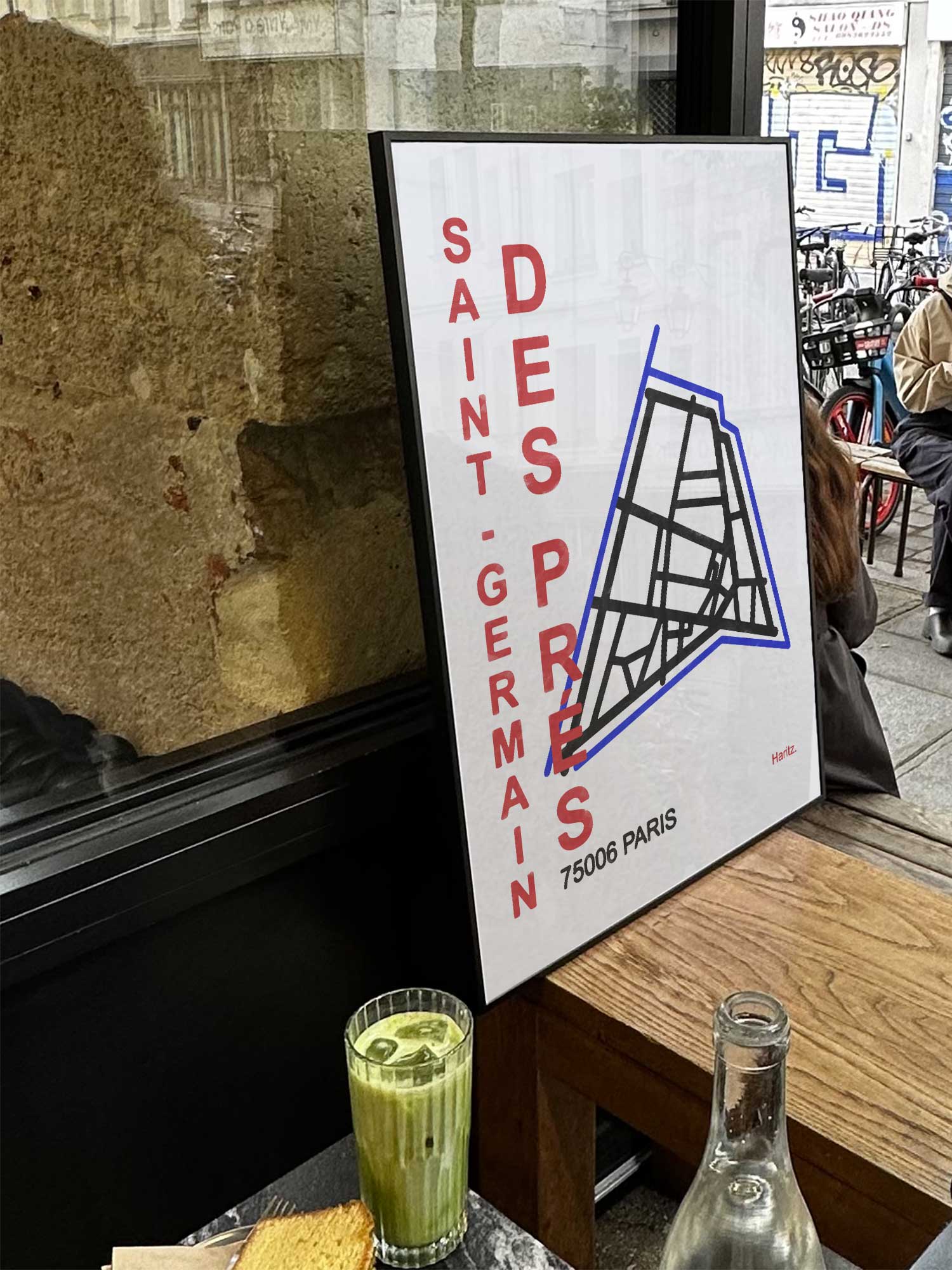 Affiche Saint Germain des Près de Haritz exposée près d'une fenêtre dans un café parisien tendance à côté d'un verre de matcha latte.