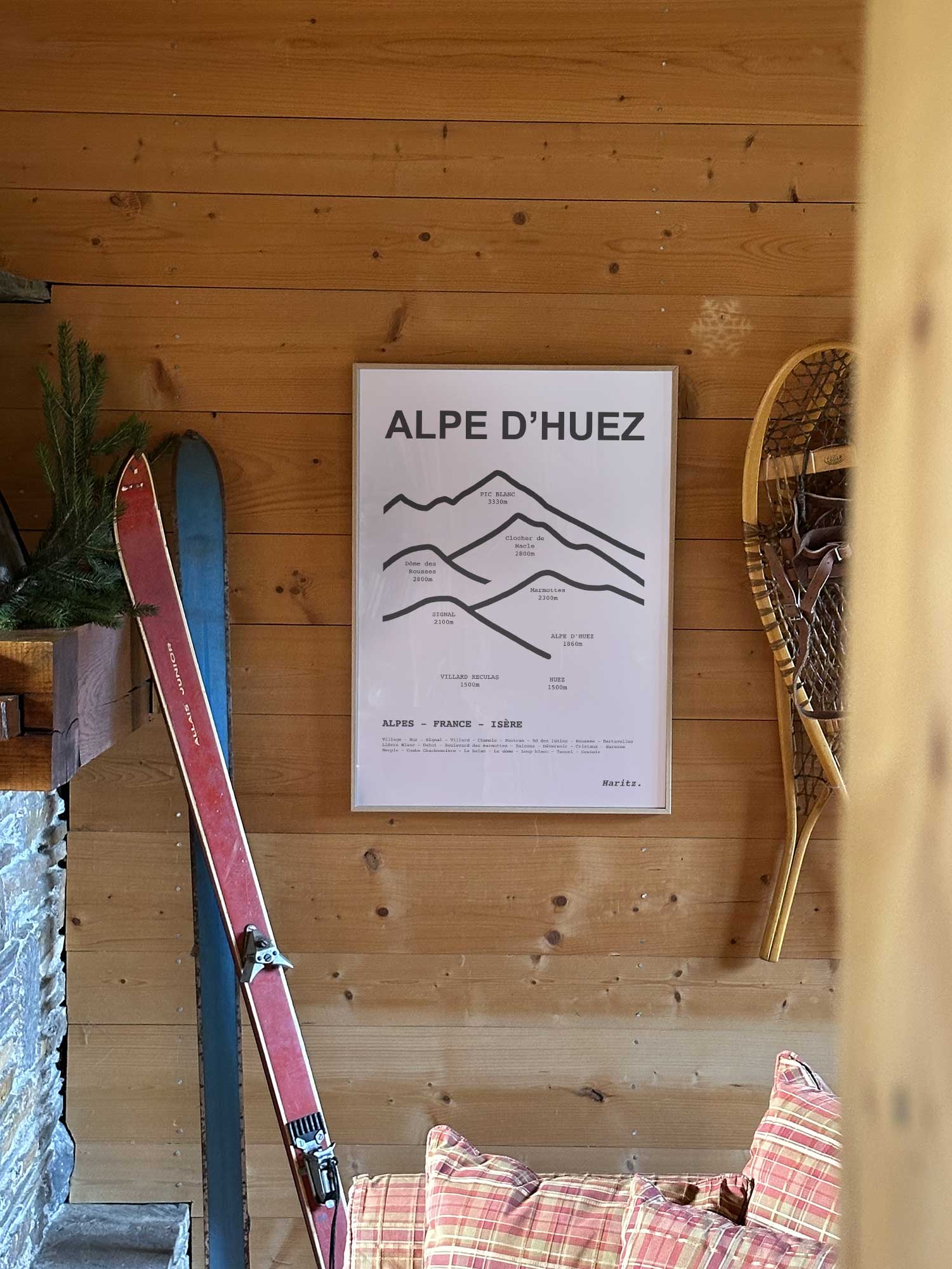 Affiche Alpe d'Huez représentant les sommets de la station, accrochée au mur d’un salon de chalet avec skis vintage et raquettes à neige, décoration montagne Haritz.